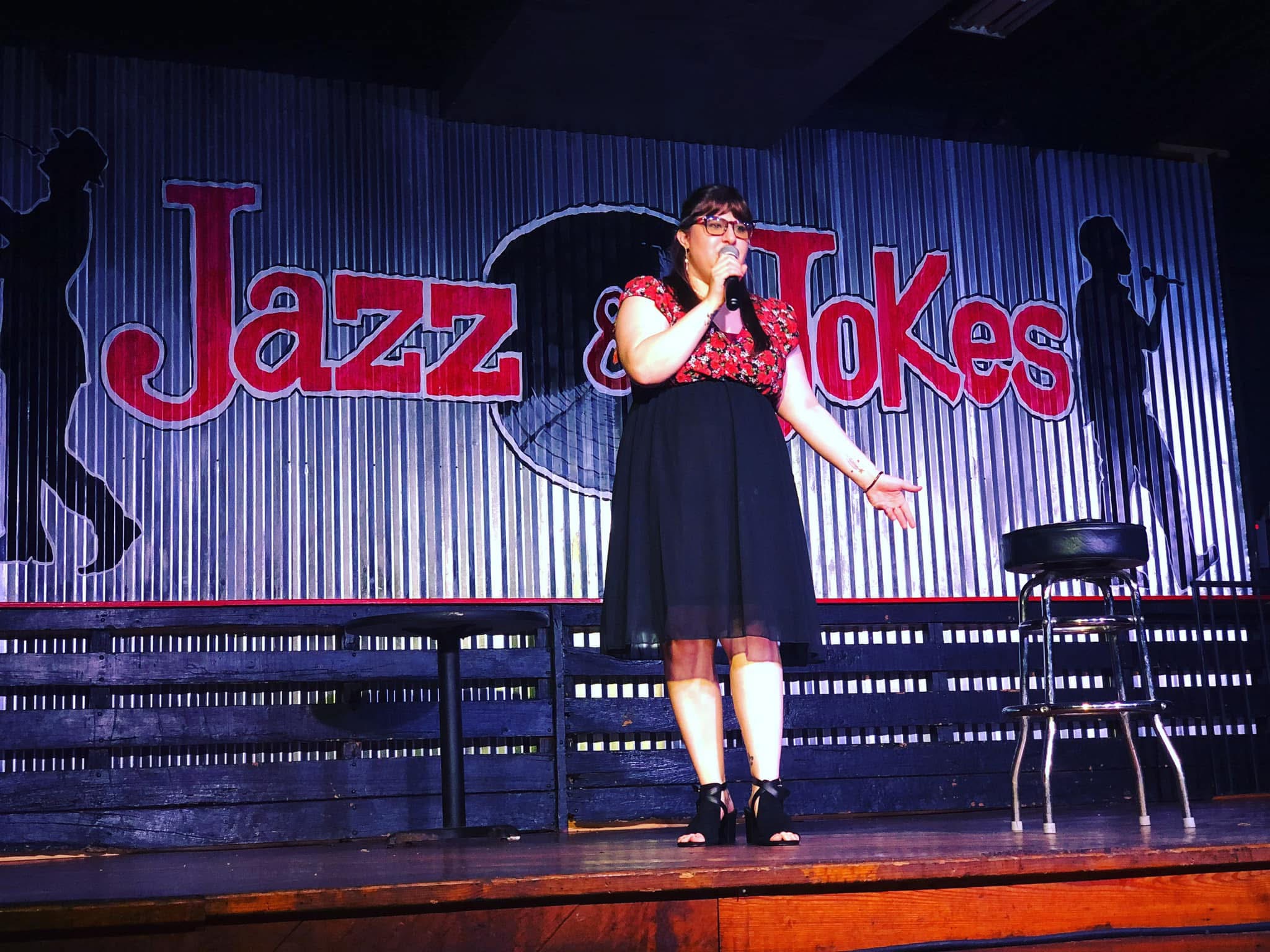 Performer on stage with a microphone, standing in front of a backdrop reading 'Jazz & Jokes,' with silhouettes of dancers.