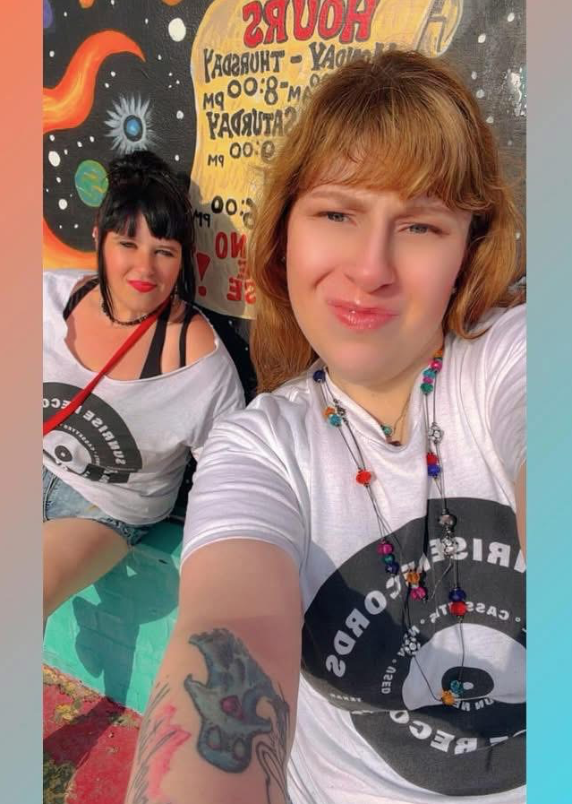 Two women taking a selfie, one with a tattoo on her arm, in front of a colorful background with text and designs.