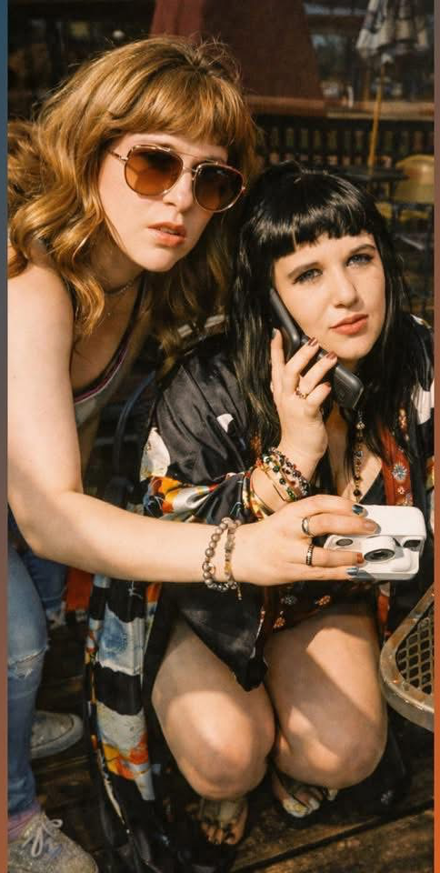 Two women with dark hair and sunglasses, one with curly hair, in a dimly lit indoor setting.