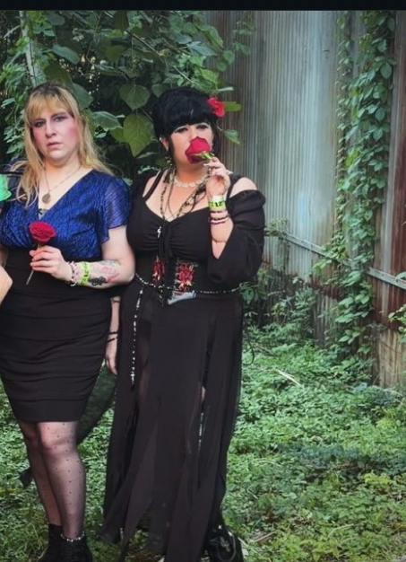 Two women standing outdoors in front of greenery and a waterfall, one holding a red rose, the other dressed as a clown.
