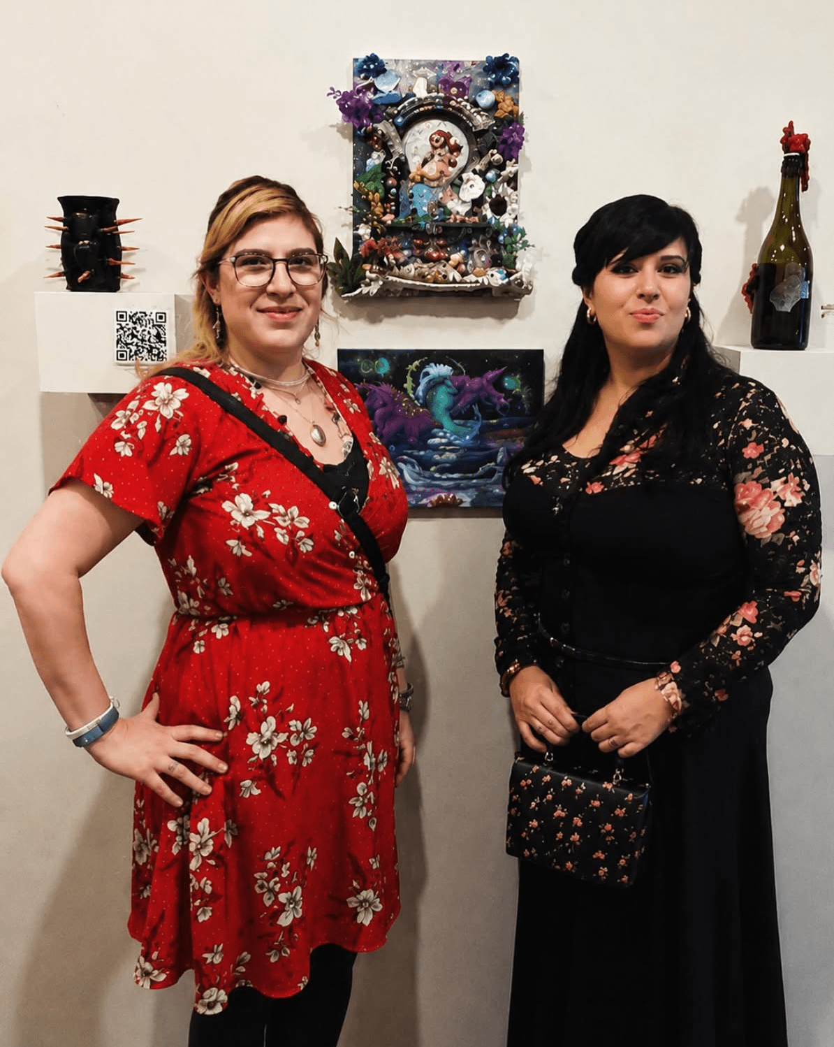 Two women stand side by side in front of a white wall with decorative items and artwork, smiling at the camera.