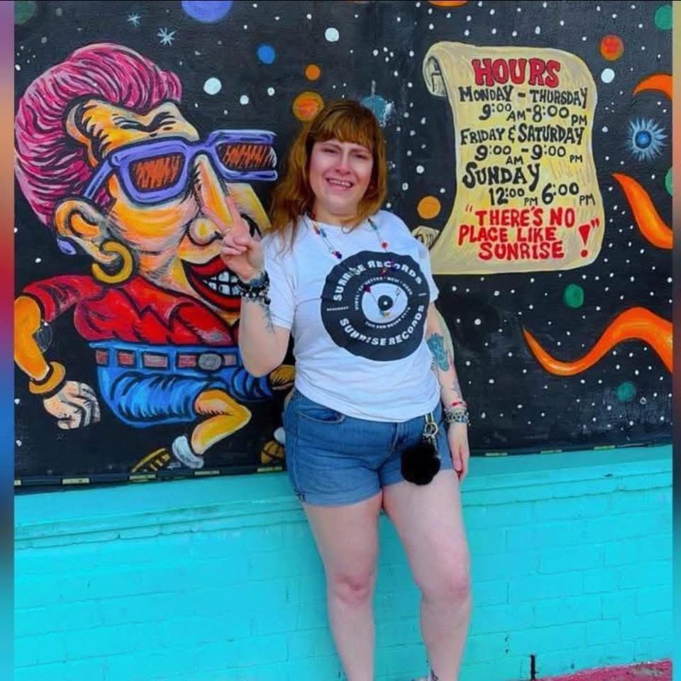Young woman with brown hair and a white T-shirt standing in front of a colorful mural, smiling and pointing upward.