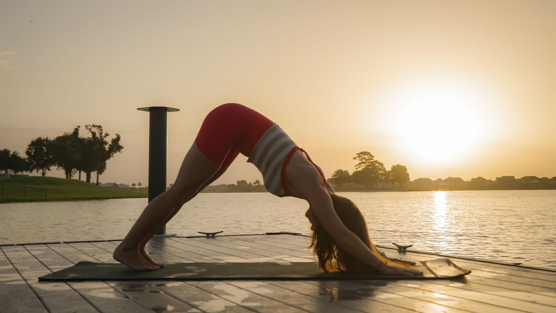 Person practicing yoga outdoors on a mat near water during sunset, in a downward dog pose.