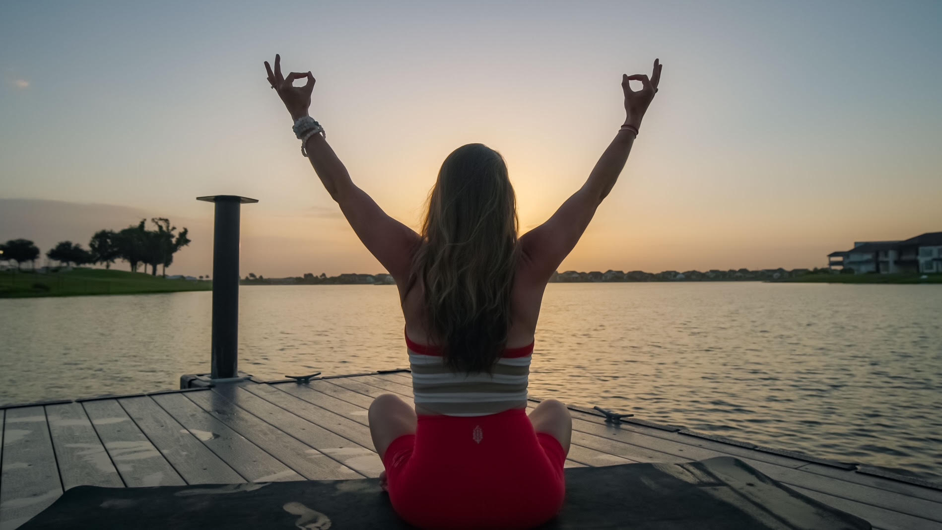 Person sitting on a dock facing water with arms raised and making peace signs, sunset in background.