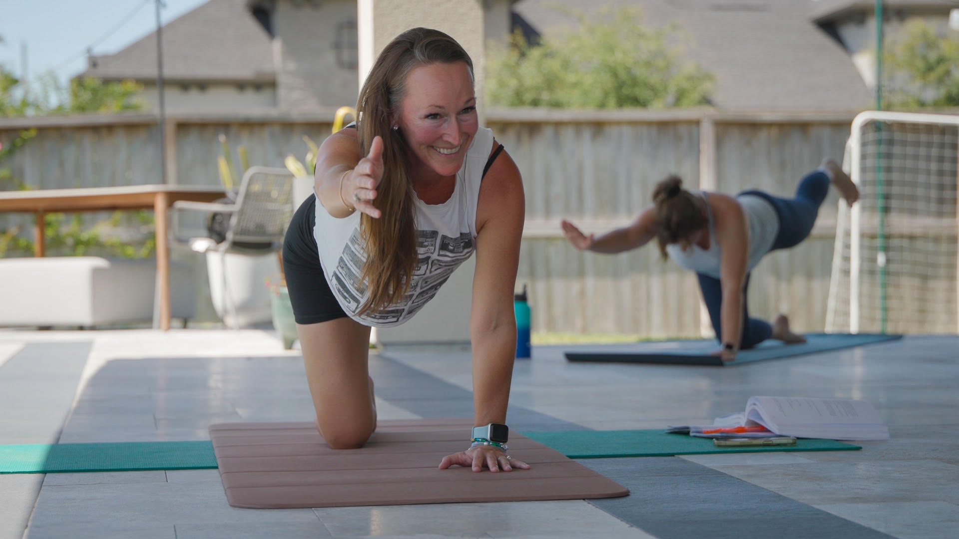 Two women practicing yoga outdoors on mats, one in foreground smiling, the other in background stretching on a mat.