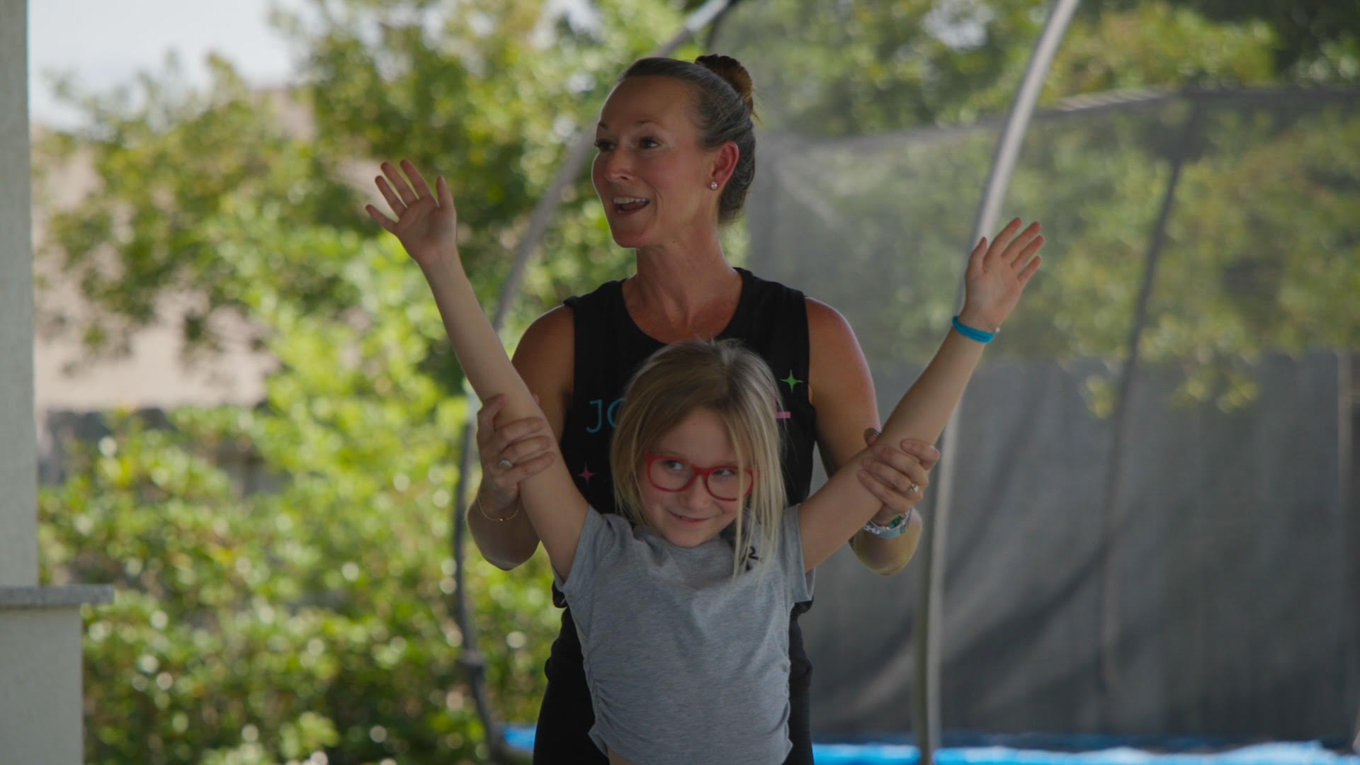 Woman and girl smiling outdoors, girl with glasses raising arms, trees and a fence in background.