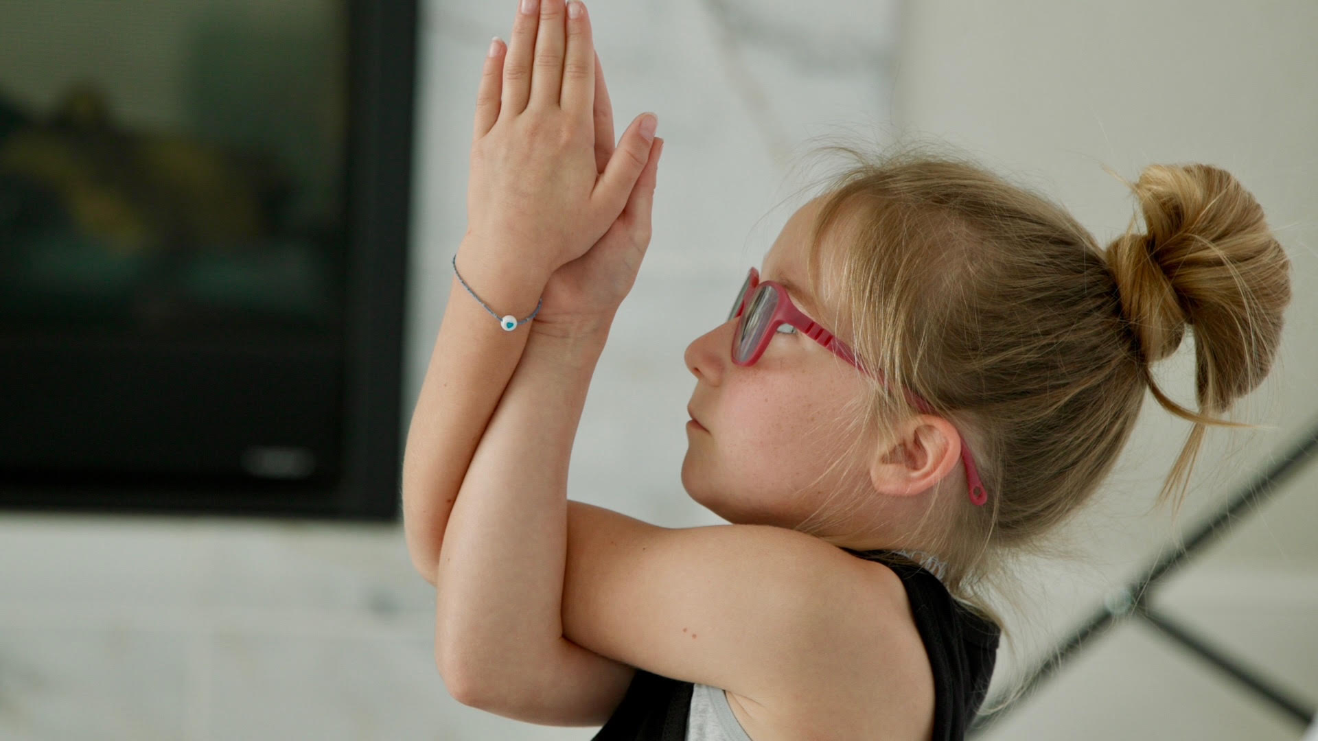 Child with glasses and a bun, hands pressed together near face, looking upward, indoors.
