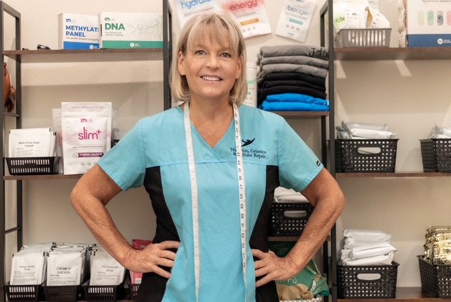 A smiling woman in medical scrubs stands in a storage room with shelves of supplies behind her.