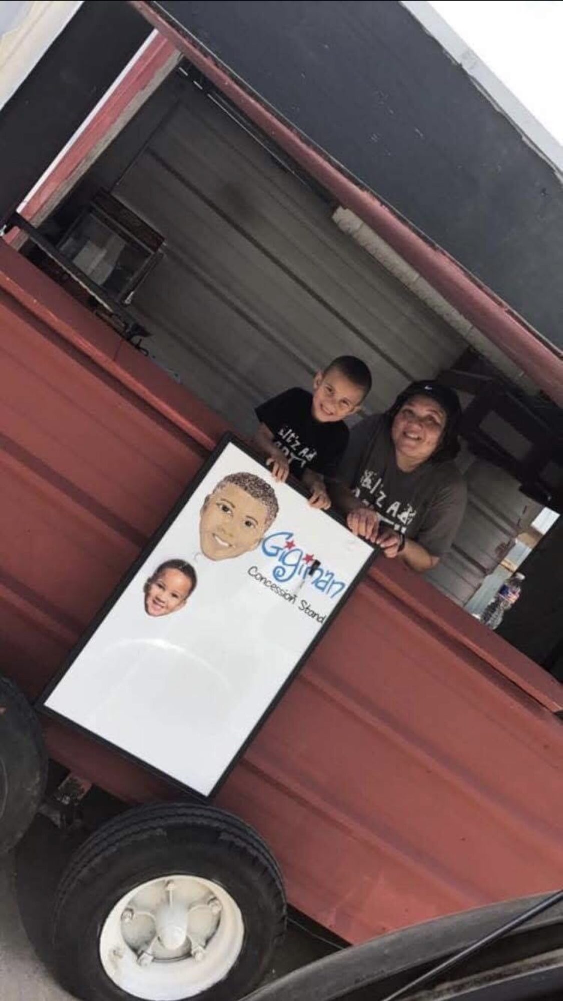 Two people stand behind a food cart with a sign featuring a child's face and the word 'Gourmet'.