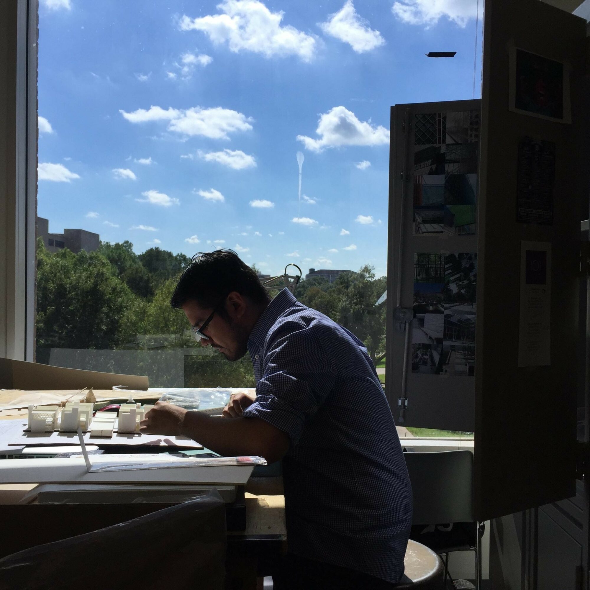Person sitting at a desk with a window showing a blue sky and clouds behind them.