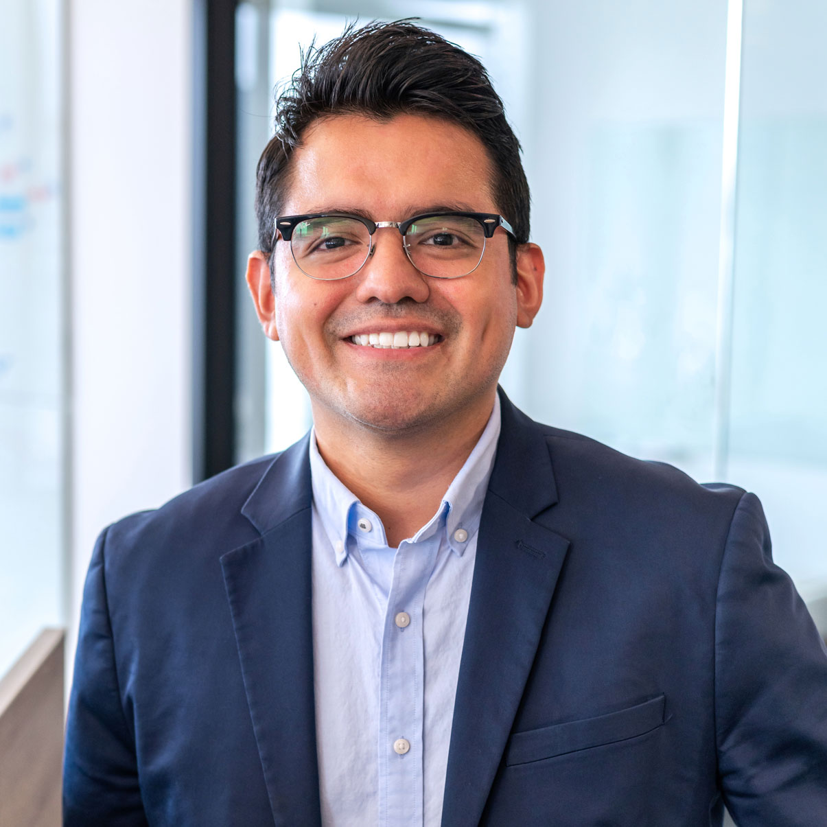 Man with glasses smiling, wearing a suit jacket and light blue shirt, in an office setting.