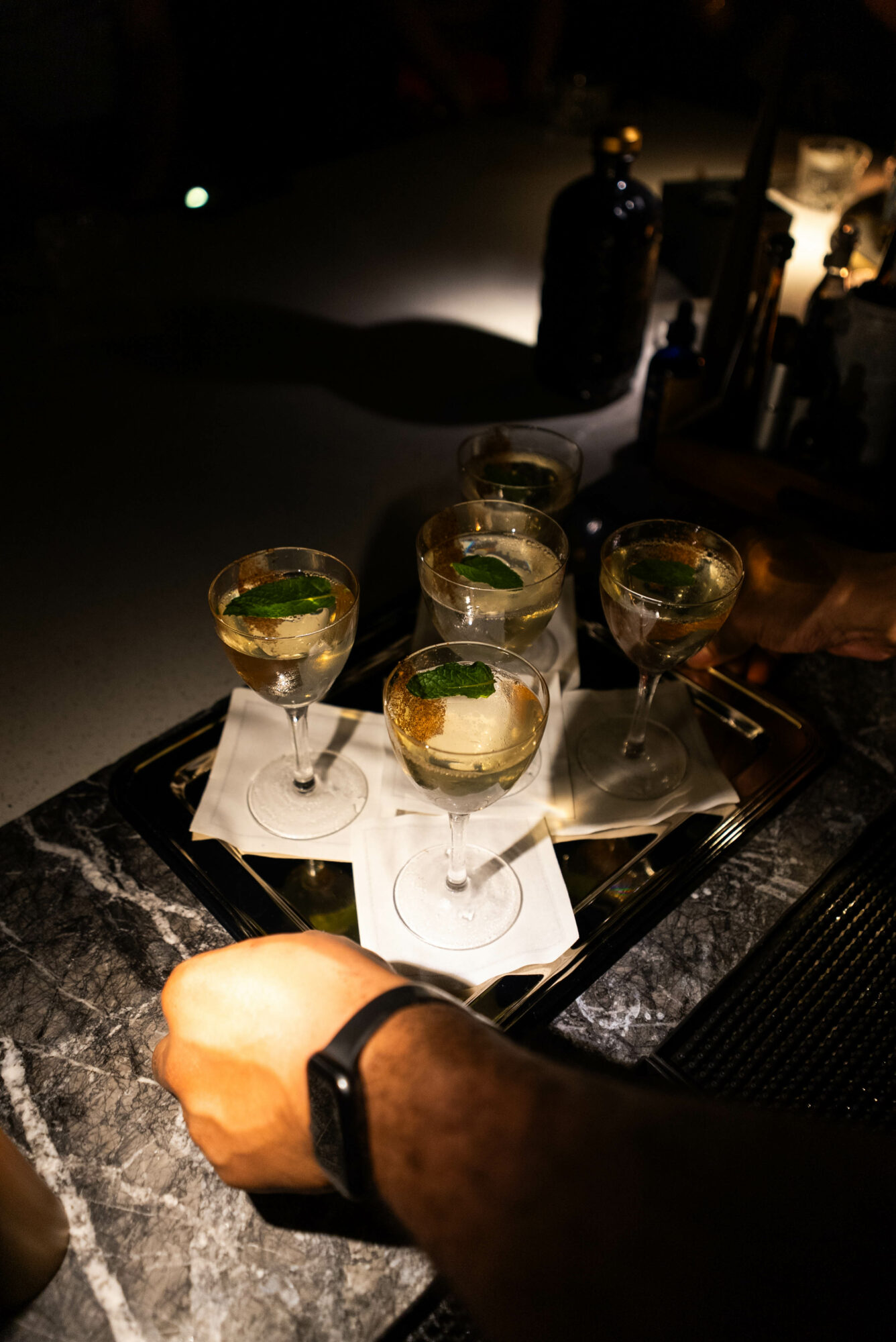 Person's hand holding a tray with five glasses of cocktails garnished with mint leaves, on a dark bar counter.