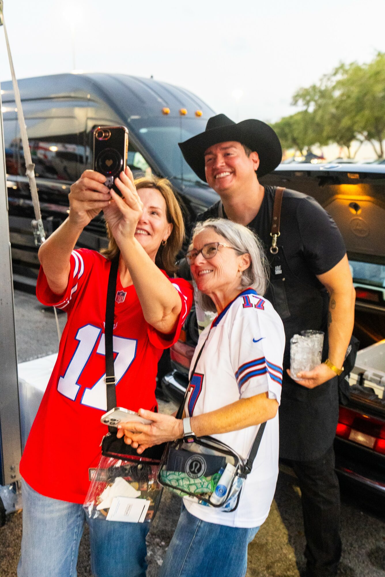 Three people smiling and taking a selfie outdoors, with a large vehicle and trees in the background.