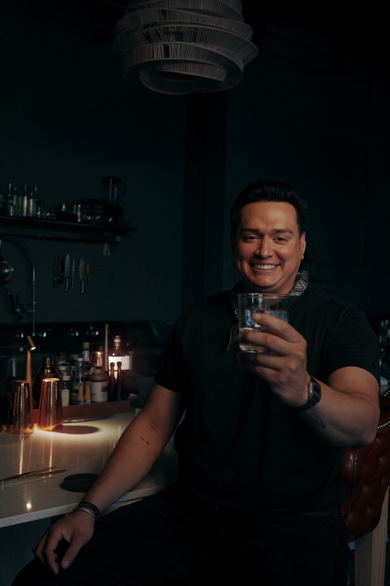 Man smiling and holding a drink in a dimly lit bar or restaurant, with bottles and glasses in the background.