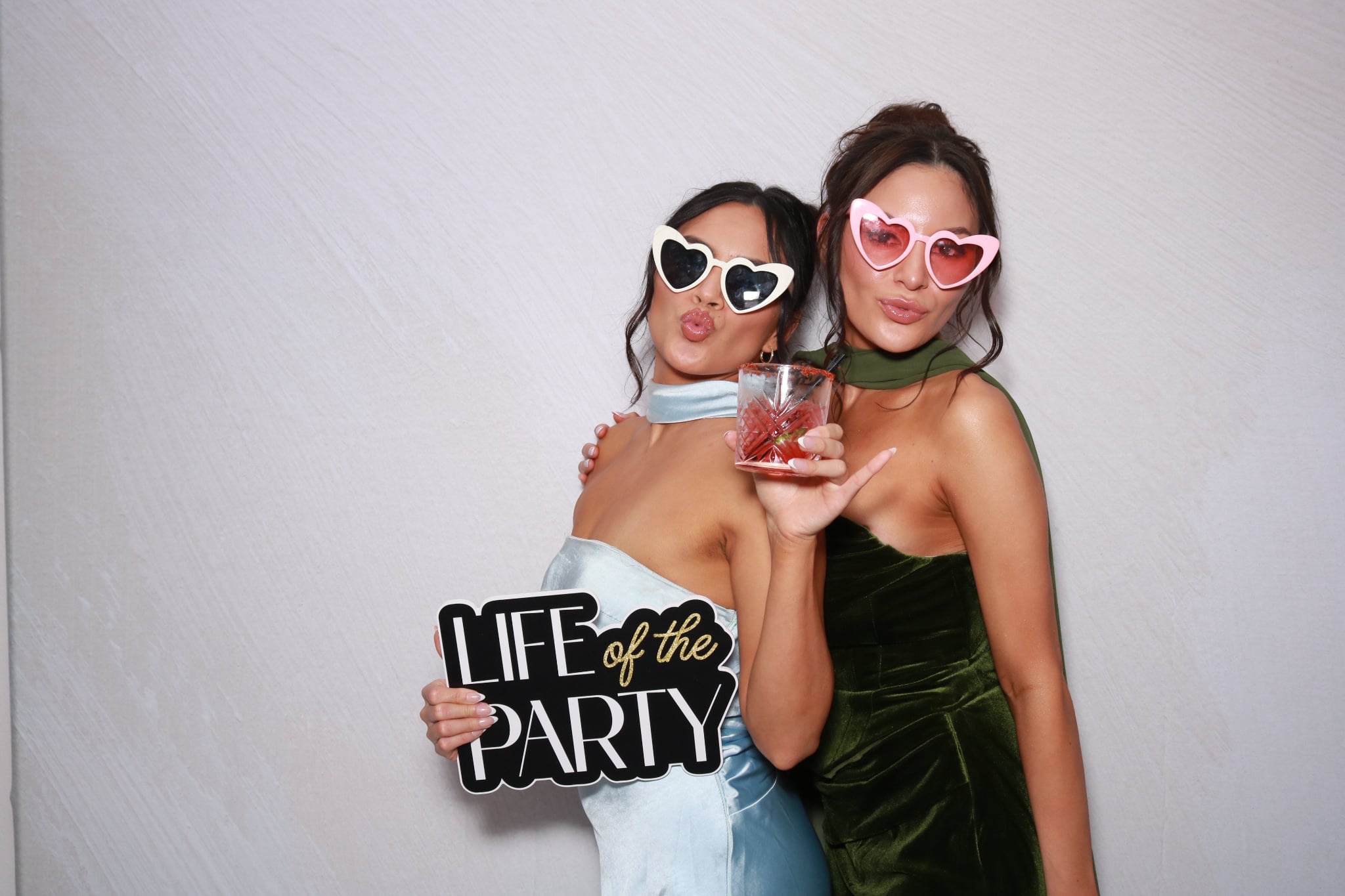 Two women wearing heart-shaped sunglasses, one holding a drink and a sign that says 'Life of the Party,' smiling together.