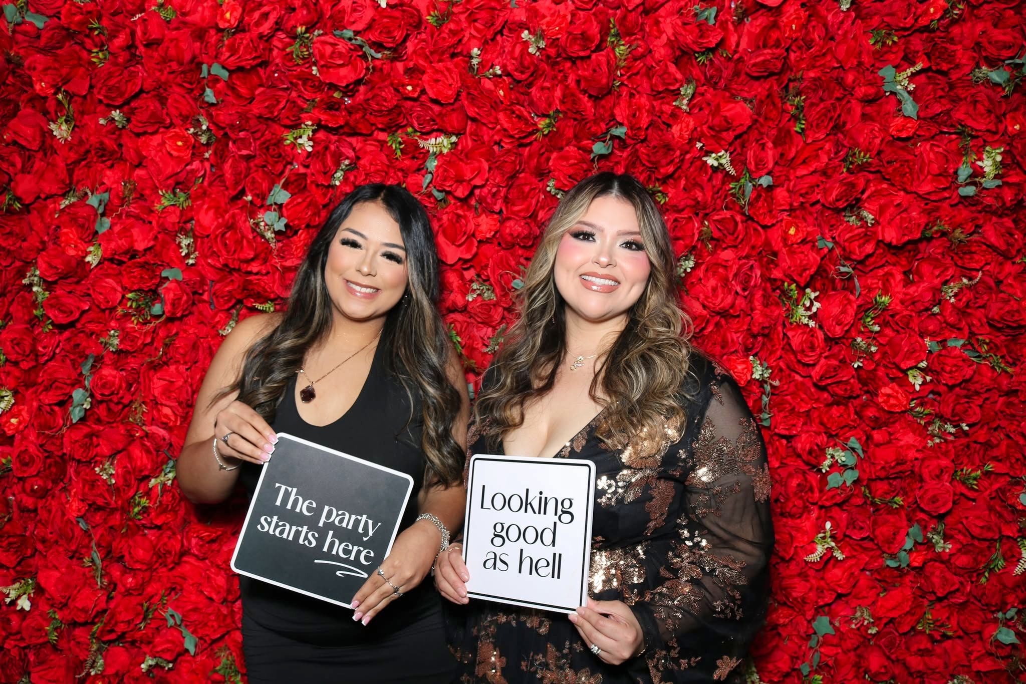Two women smiling and holding signs in front of a red flower wall, one with a black dress and the other with a lace top.