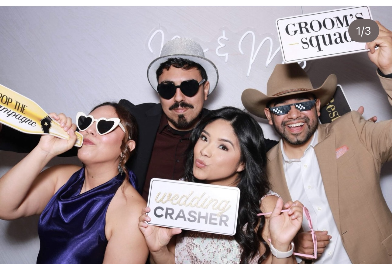 Group of four people at a wedding photo booth holding signs, wearing sunglasses and hats, smiling and posing.