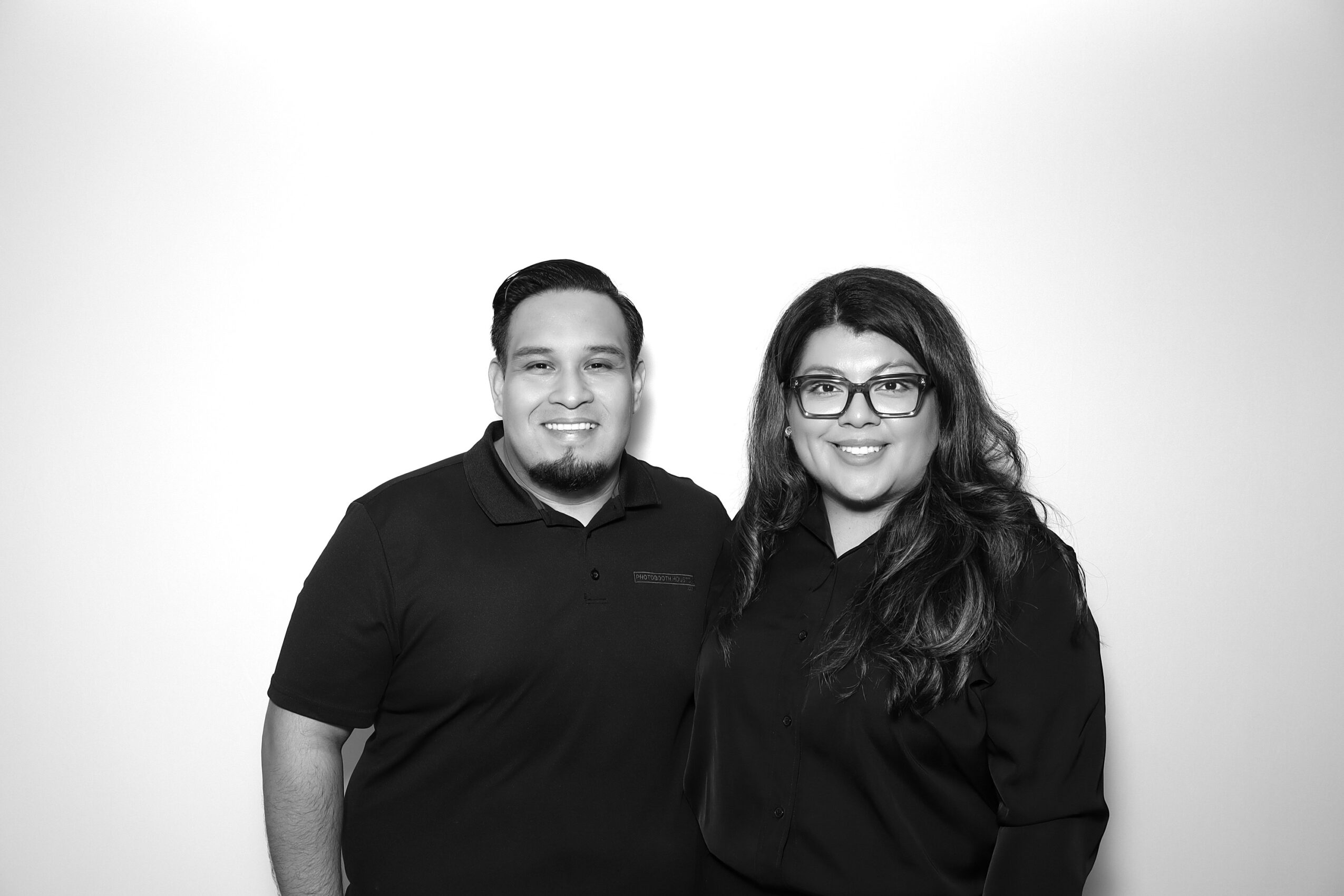 Two people standing side by side, smiling, against a plain background, one wearing glasses and the other with short hair.