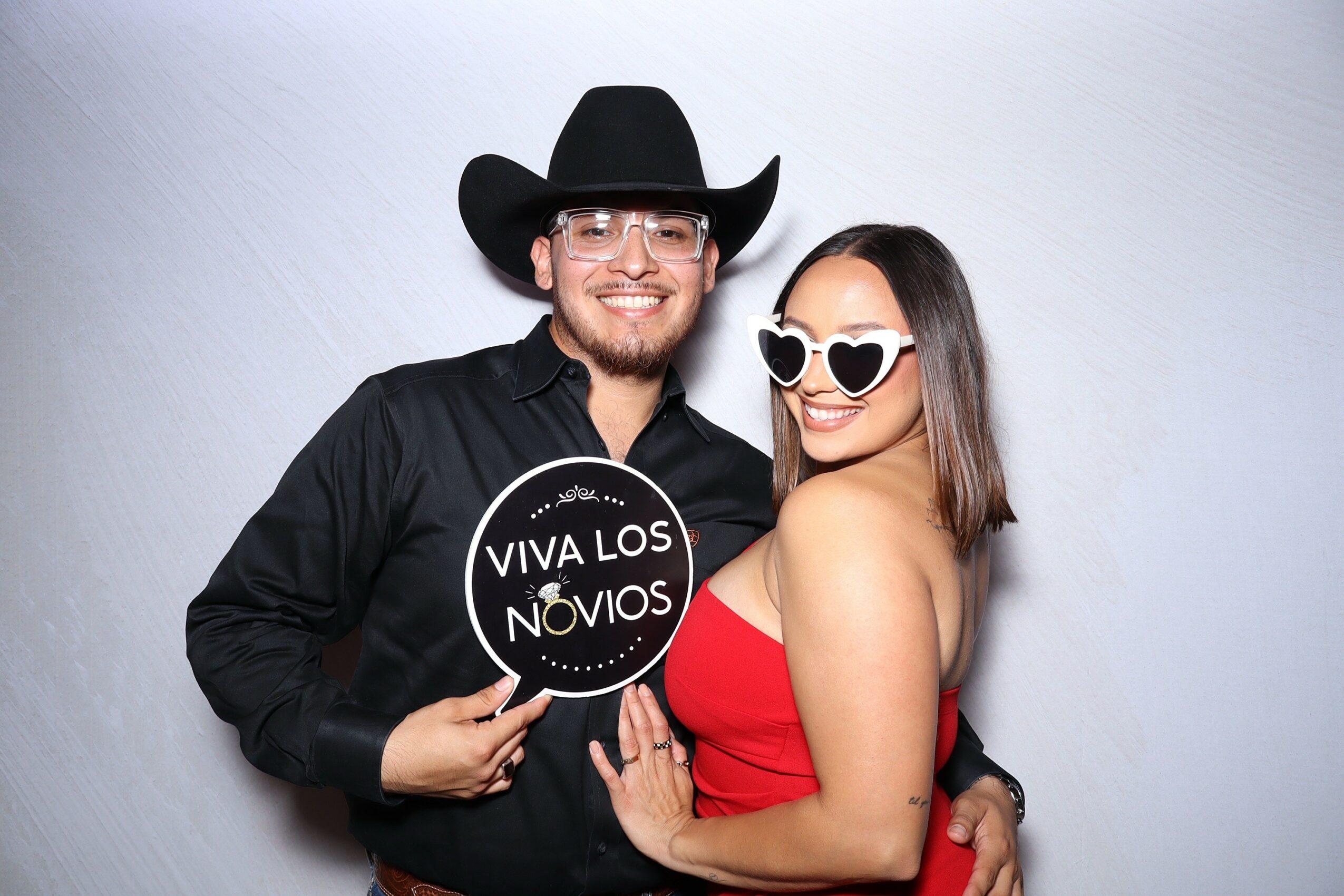 A smiling couple poses with props, the man in a cowboy hat and glasses, the woman in heart-shaped sunglasses and a red dress.