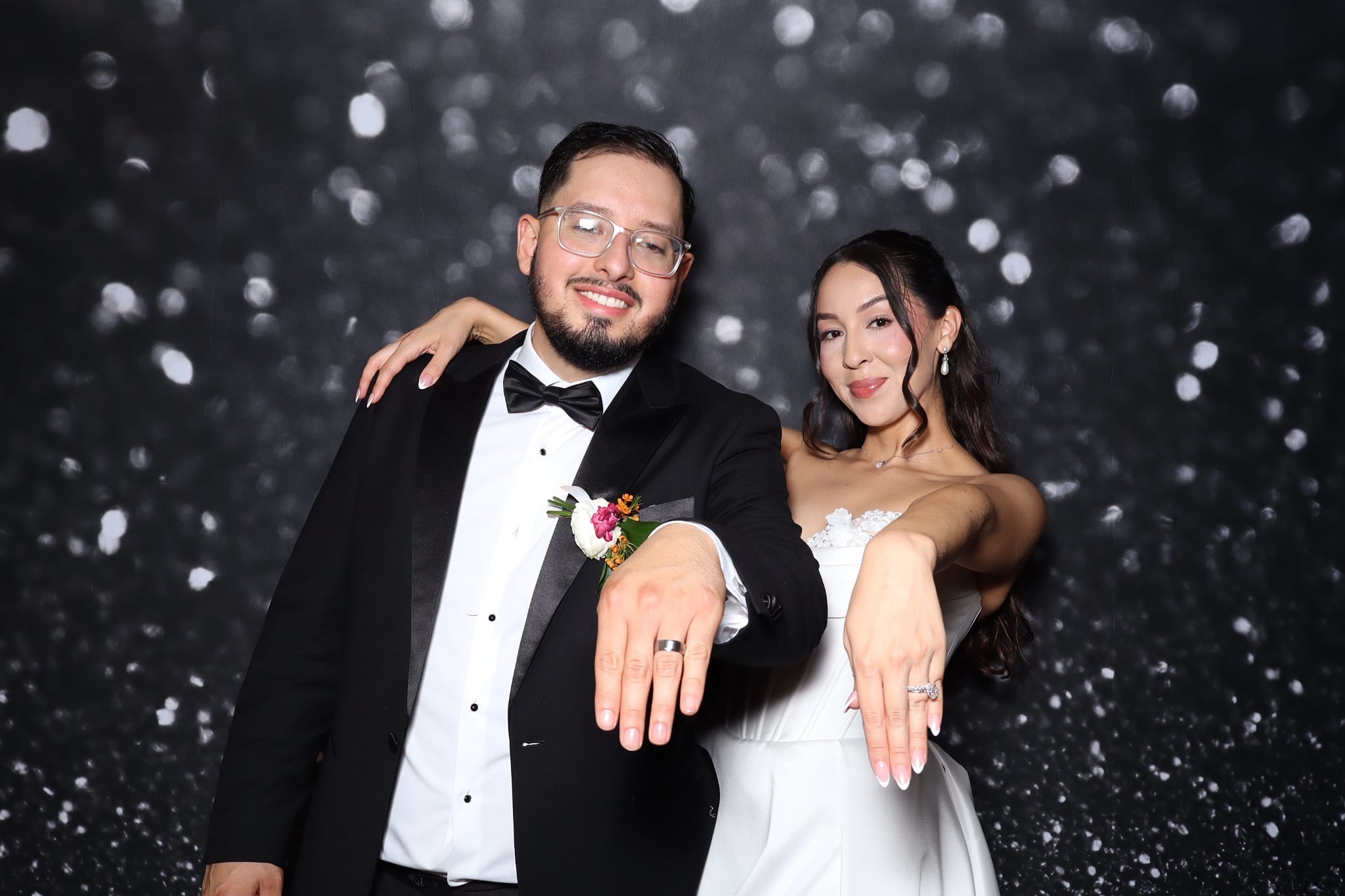 A smiling couple in formal attire, the man in a tuxedo and the woman in a white dress, pose with hands extended.