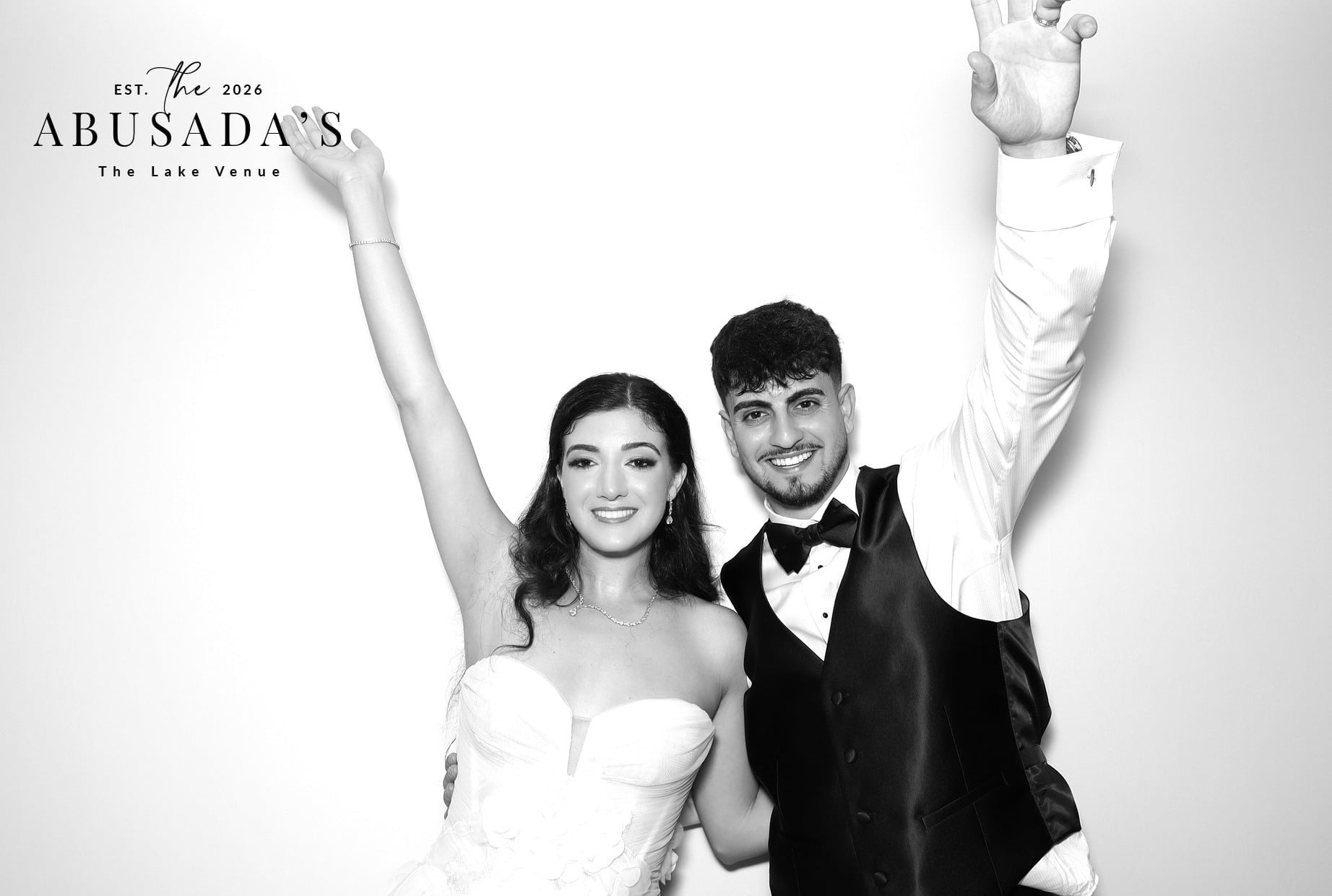 A smiling couple in formal attire, the woman in a strapless dress and the man in a tuxedo, pose with arms raised.