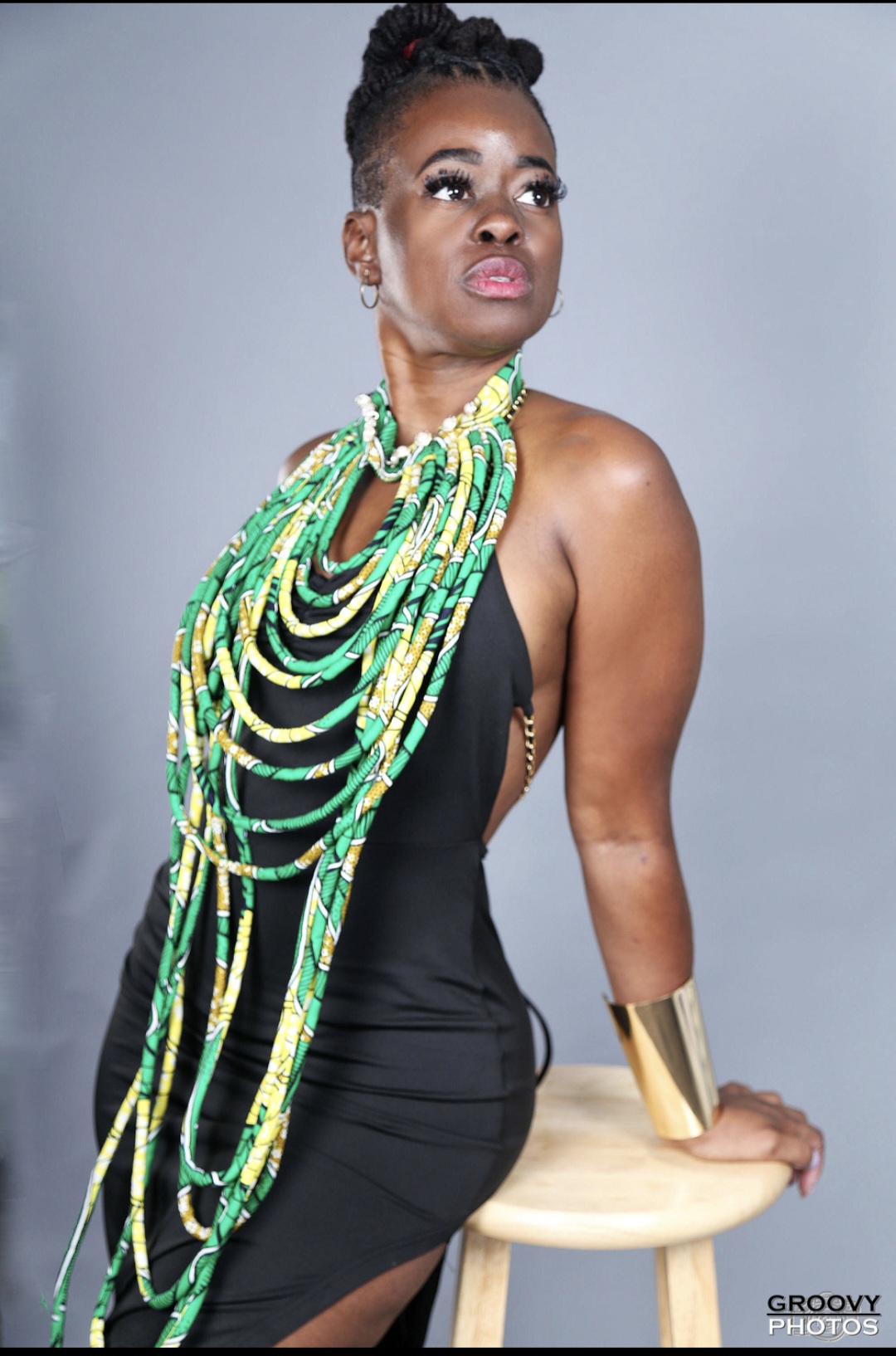 Woman with elaborate green and yellow beaded necklace, wearing a black dress, sitting on a stool against a gray background.