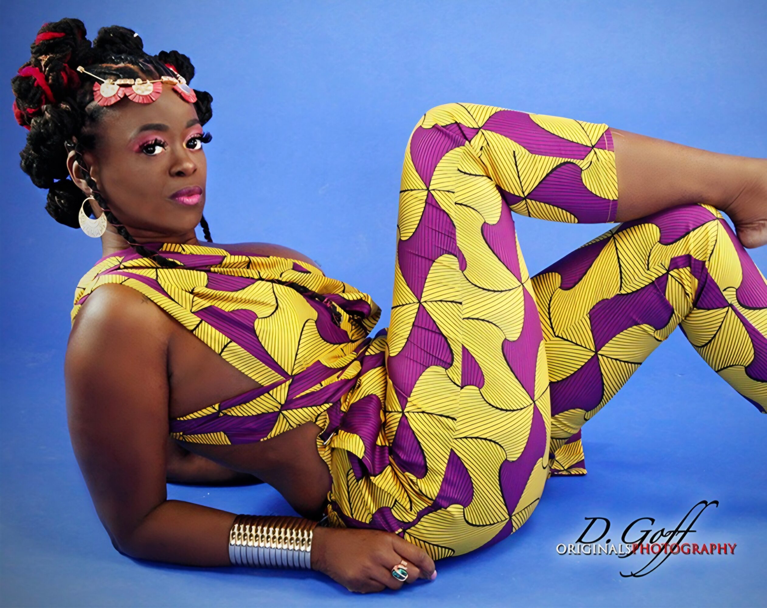 Woman in colorful patterned outfit lying on blue background, with styled hair and accessories.