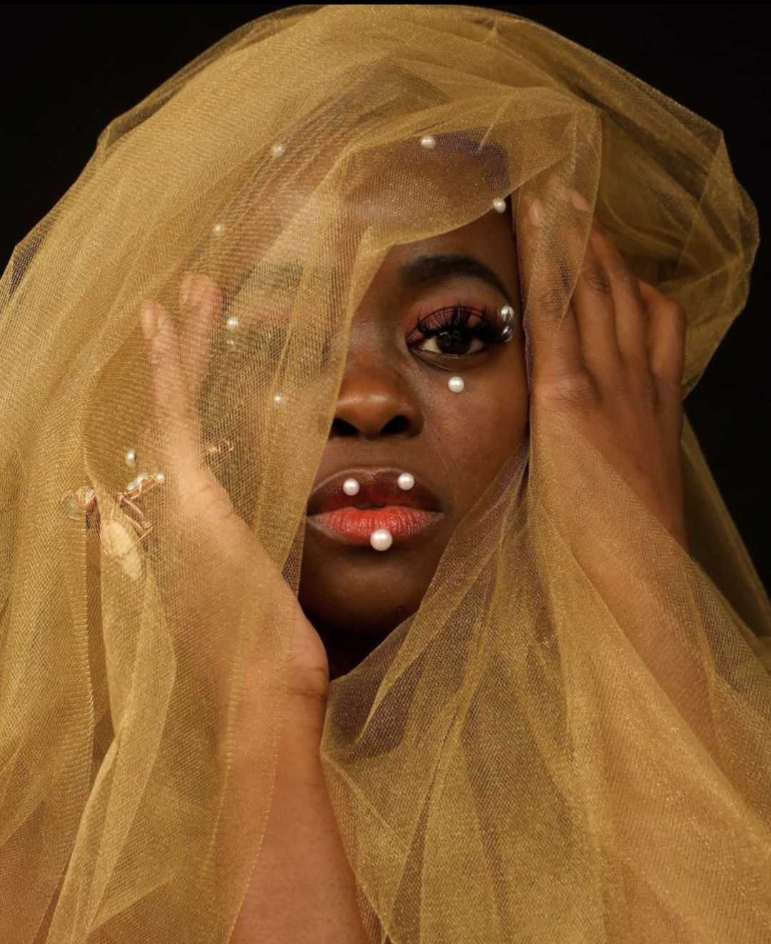 Person with gold veil covering face, holding veil with hands, dark background, makeup, and jewelry visible.