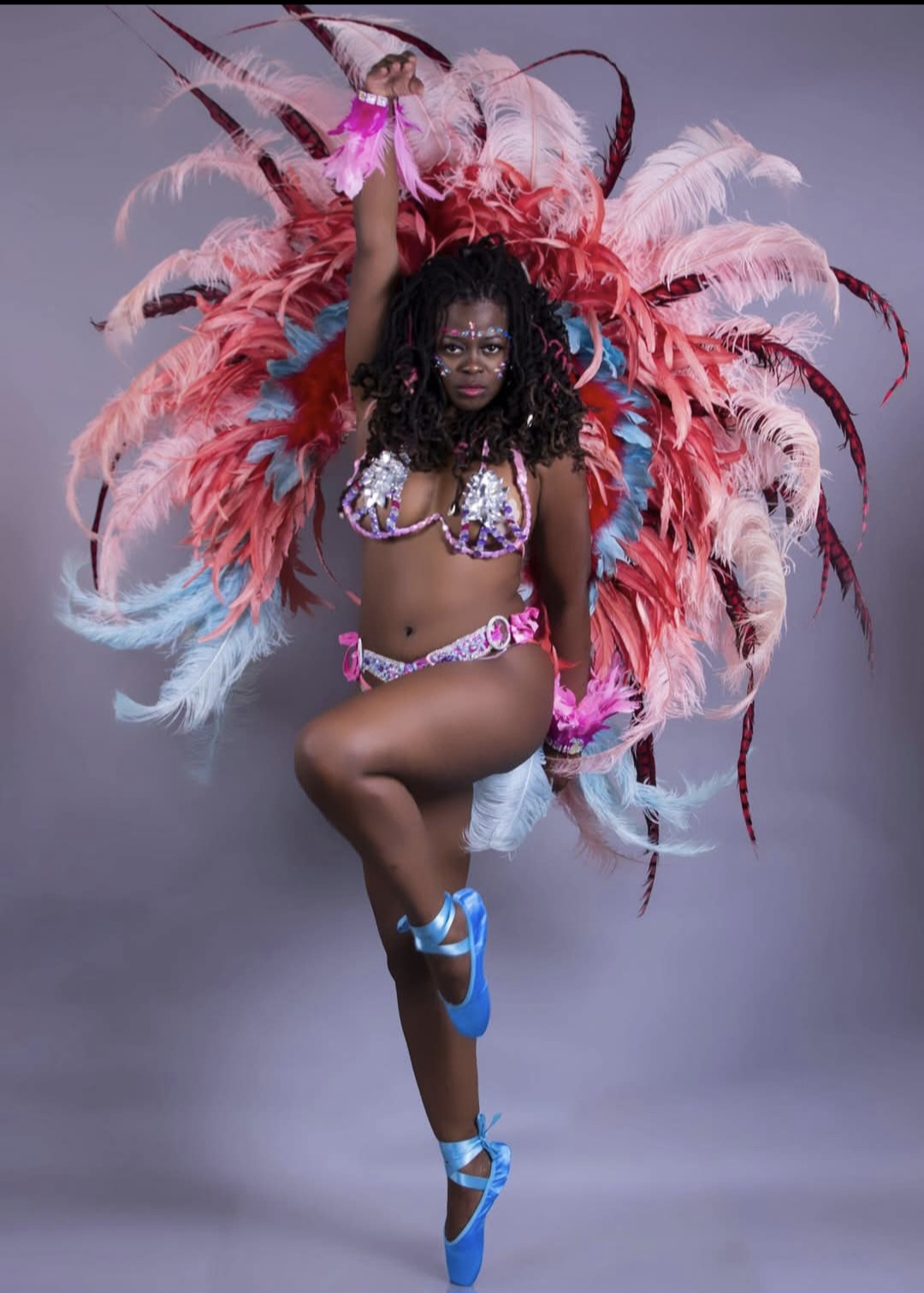 Person in colorful feathered costume with pink, red, and blue feathers, standing on one leg, arms raised, against a plain background.