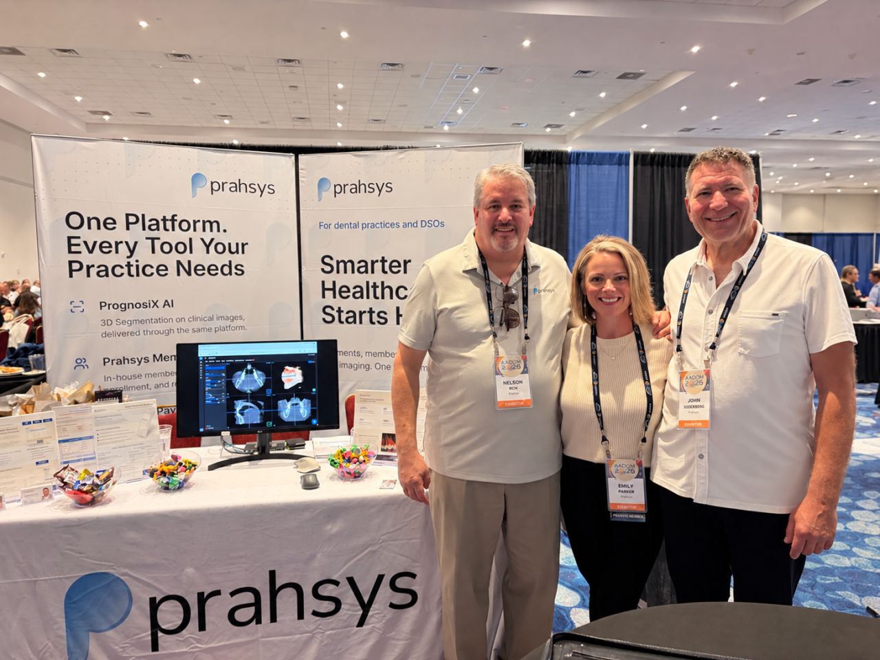 Three people standing together in front of a Prahys booth at a conference, smiling, with banners and a monitor behind them.