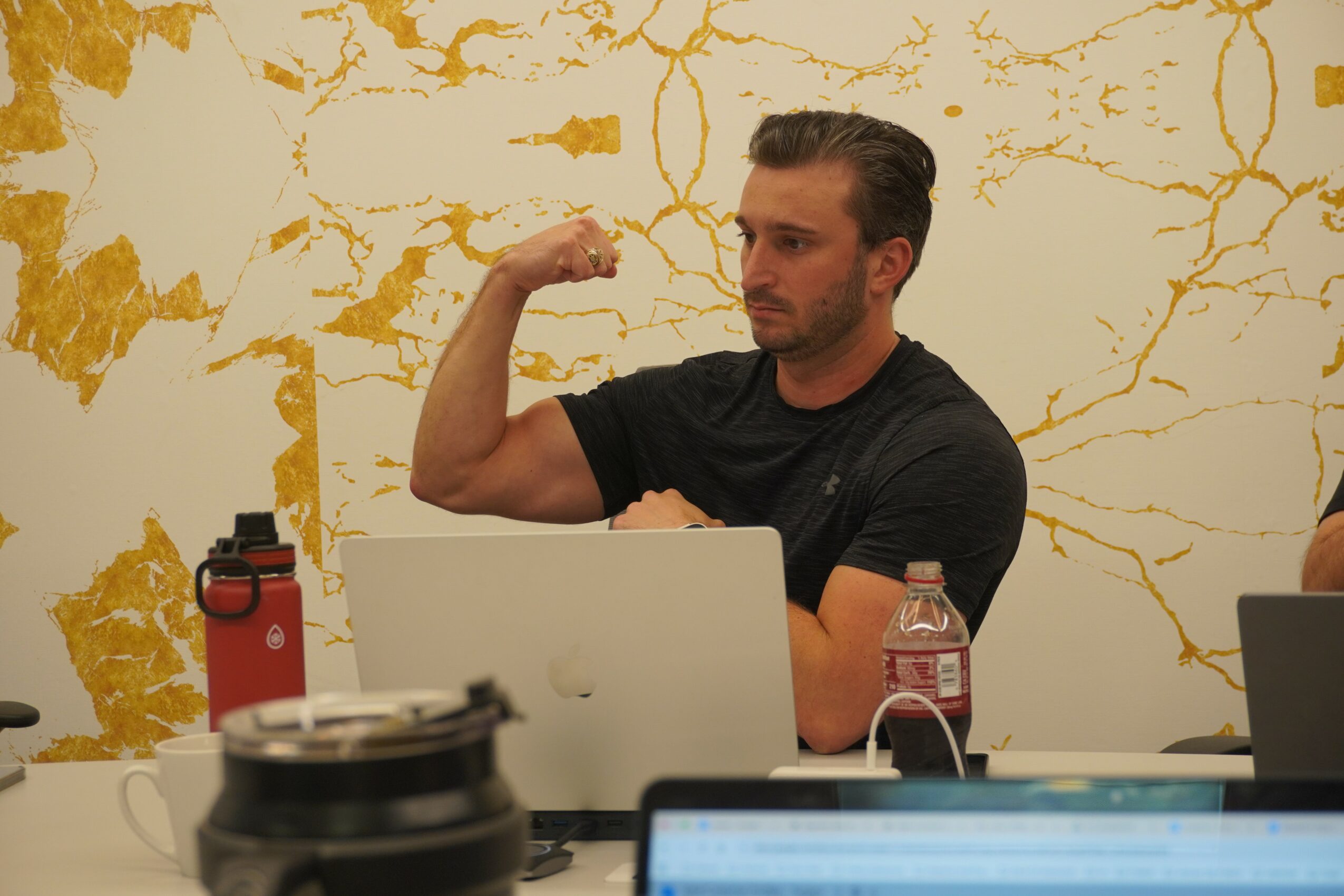 Man with dark hair and beard flexing arm muscle in front of a laptop, with a water bottle and a red thermos nearby.