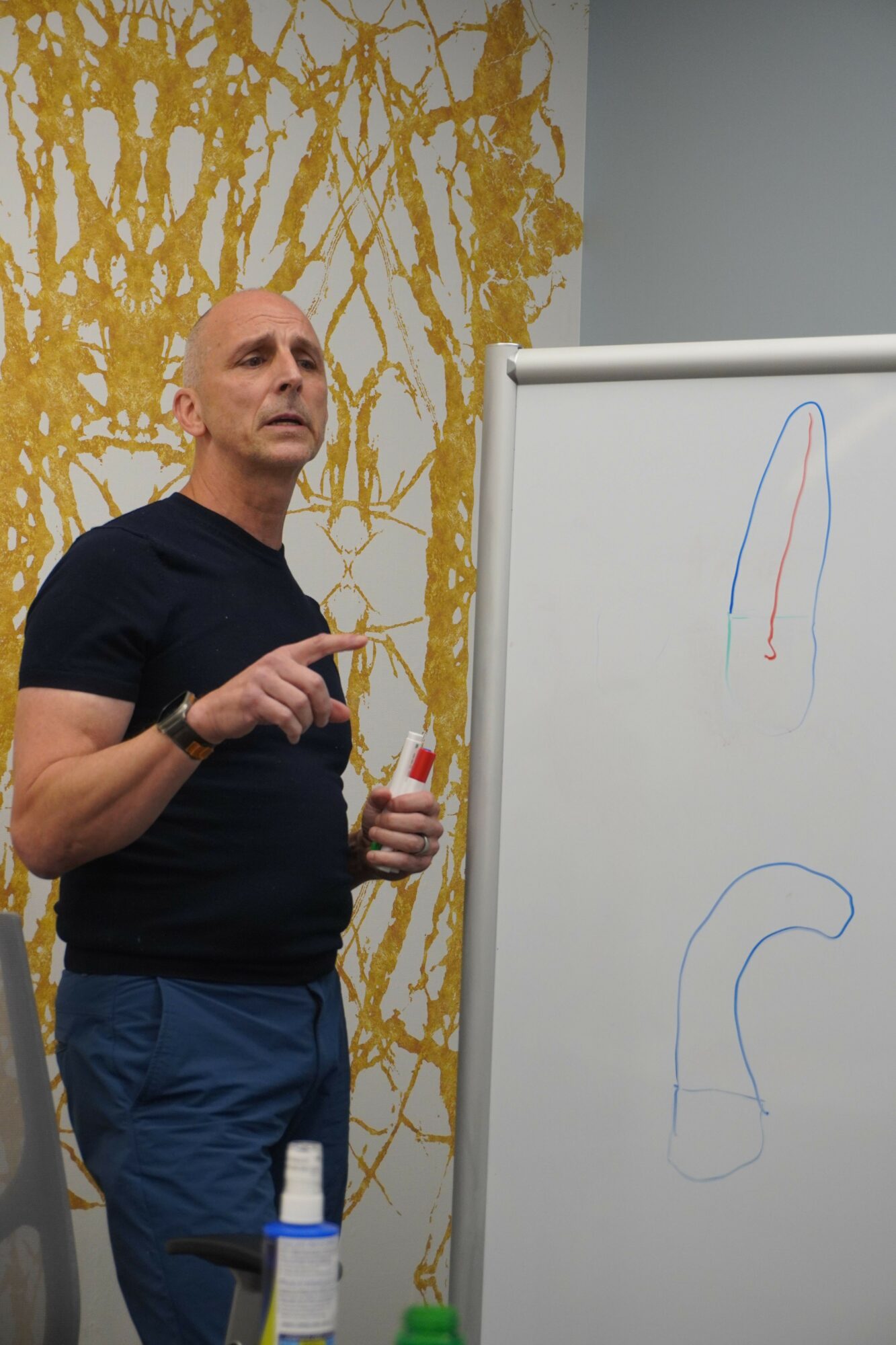 Man standing next to a whiteboard with colorful lines, in front of a gold abstract wall art, holding a marker.