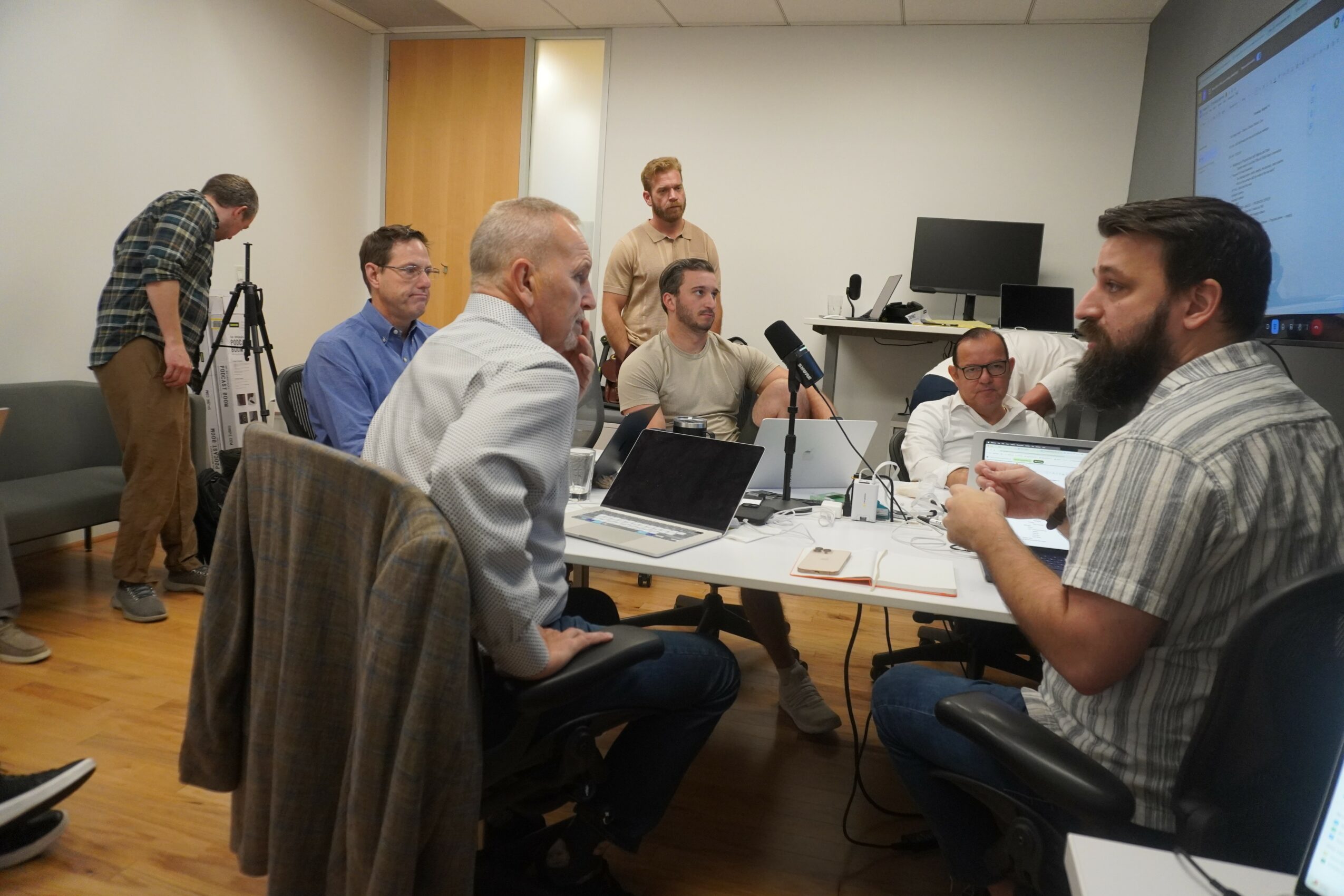 Group of people in a meeting room, some seated at a table with laptops, one person standing near a whiteboard, others observing.