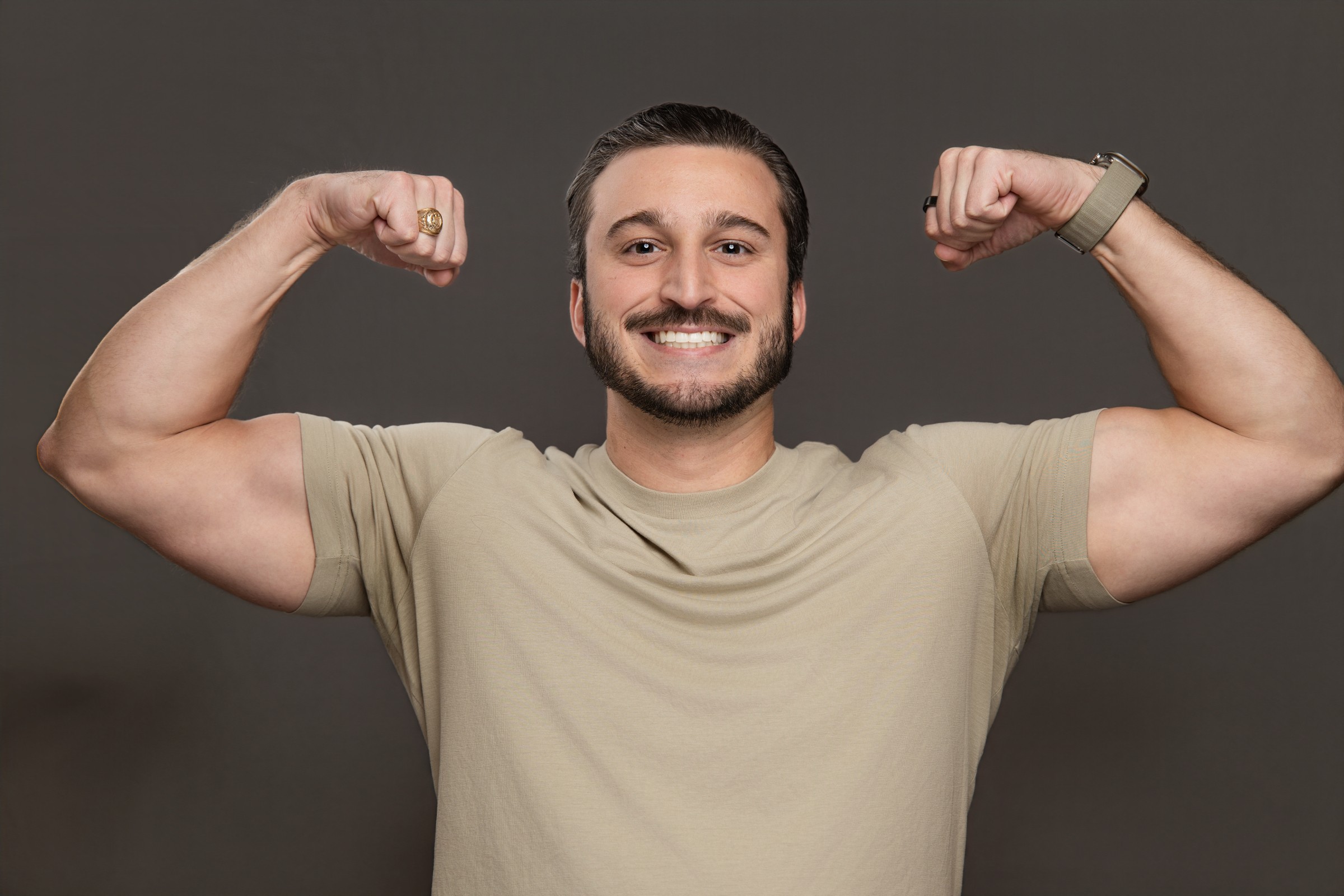 Man smiling and flexing his biceps with arms raised, wearing a beige t-shirt, against a dark background.