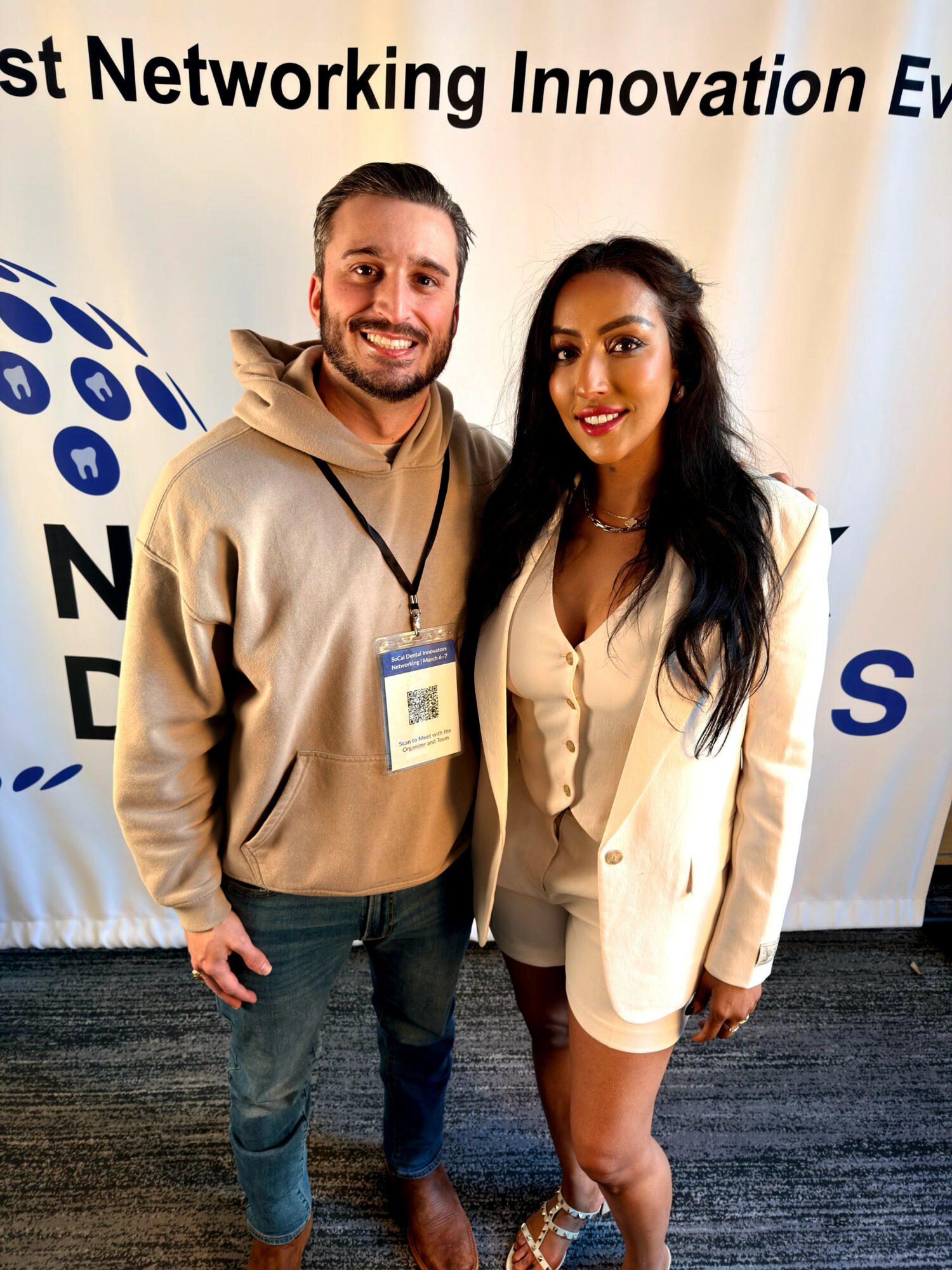 Two people standing in front of a banner, smiling, with one wearing a beige hoodie and the other in a beige blazer.