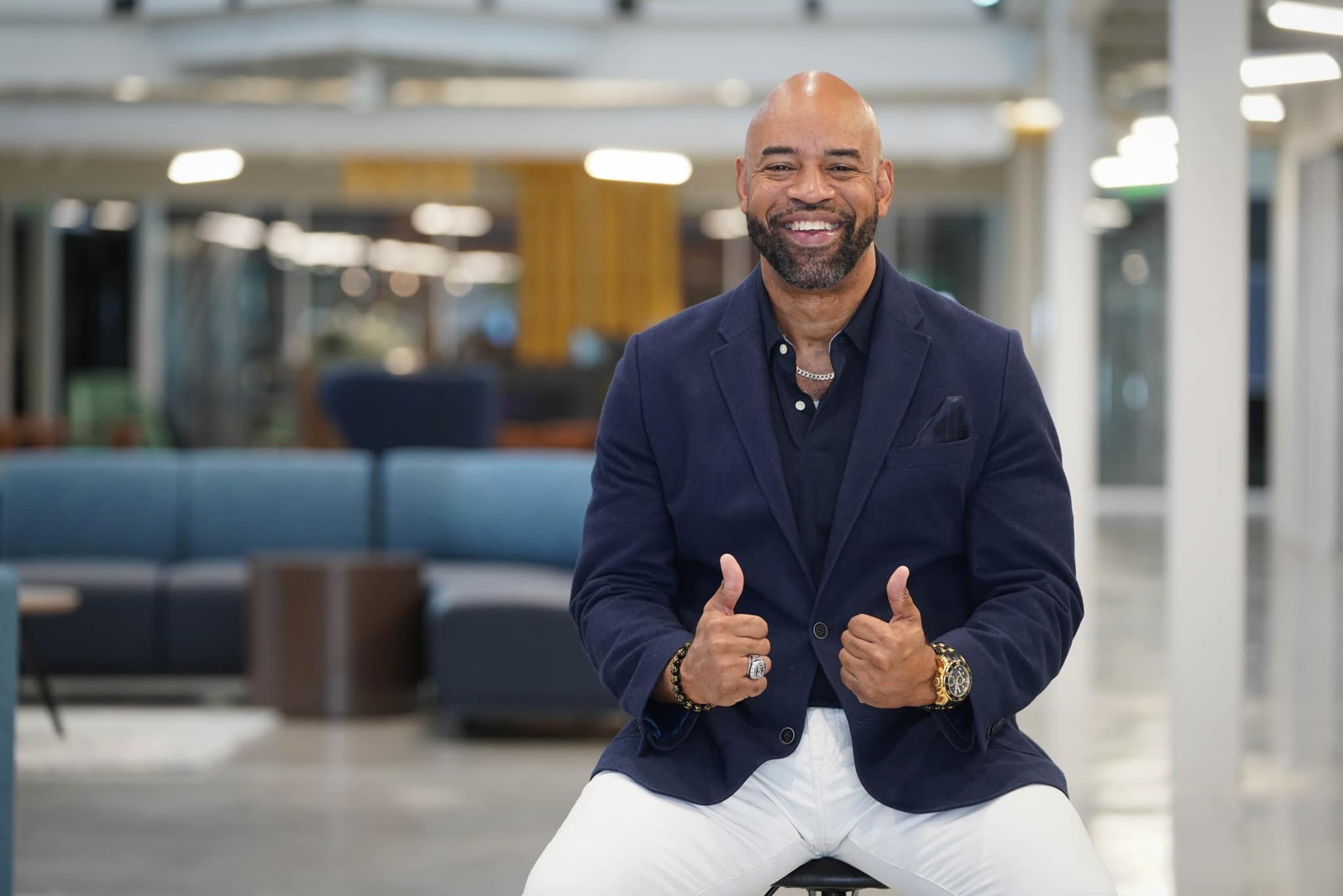 Smiling man in a dark blazer and white pants giving thumbs up in a modern indoor space.