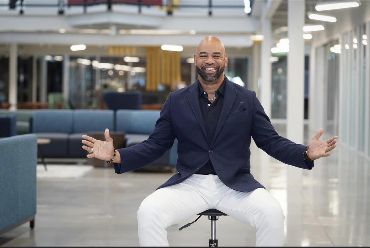 Man sitting on a stool with arms open in a modern, brightly lit indoor space, smiling broadly.