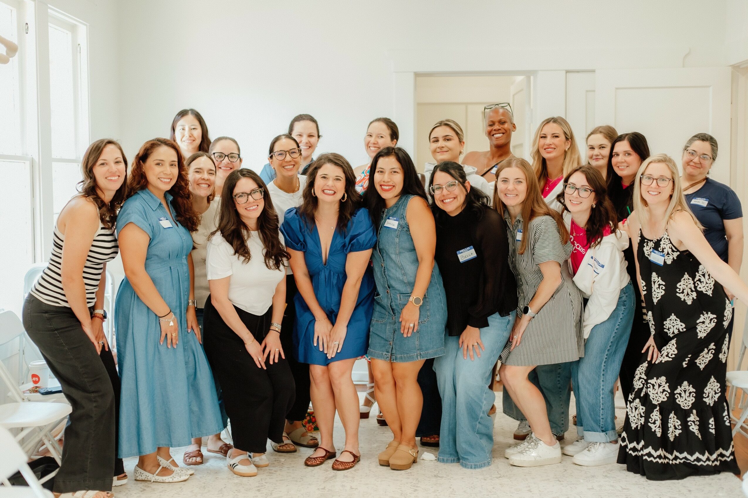Group of women and girls smiling indoors, some wearing blue dresses or casual clothing, in a bright room with white walls.