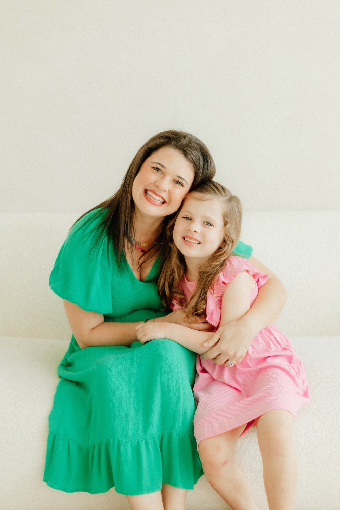 Woman and girl sitting together, smiling, woman in green dress, girl in pink dress, hugging, white background
