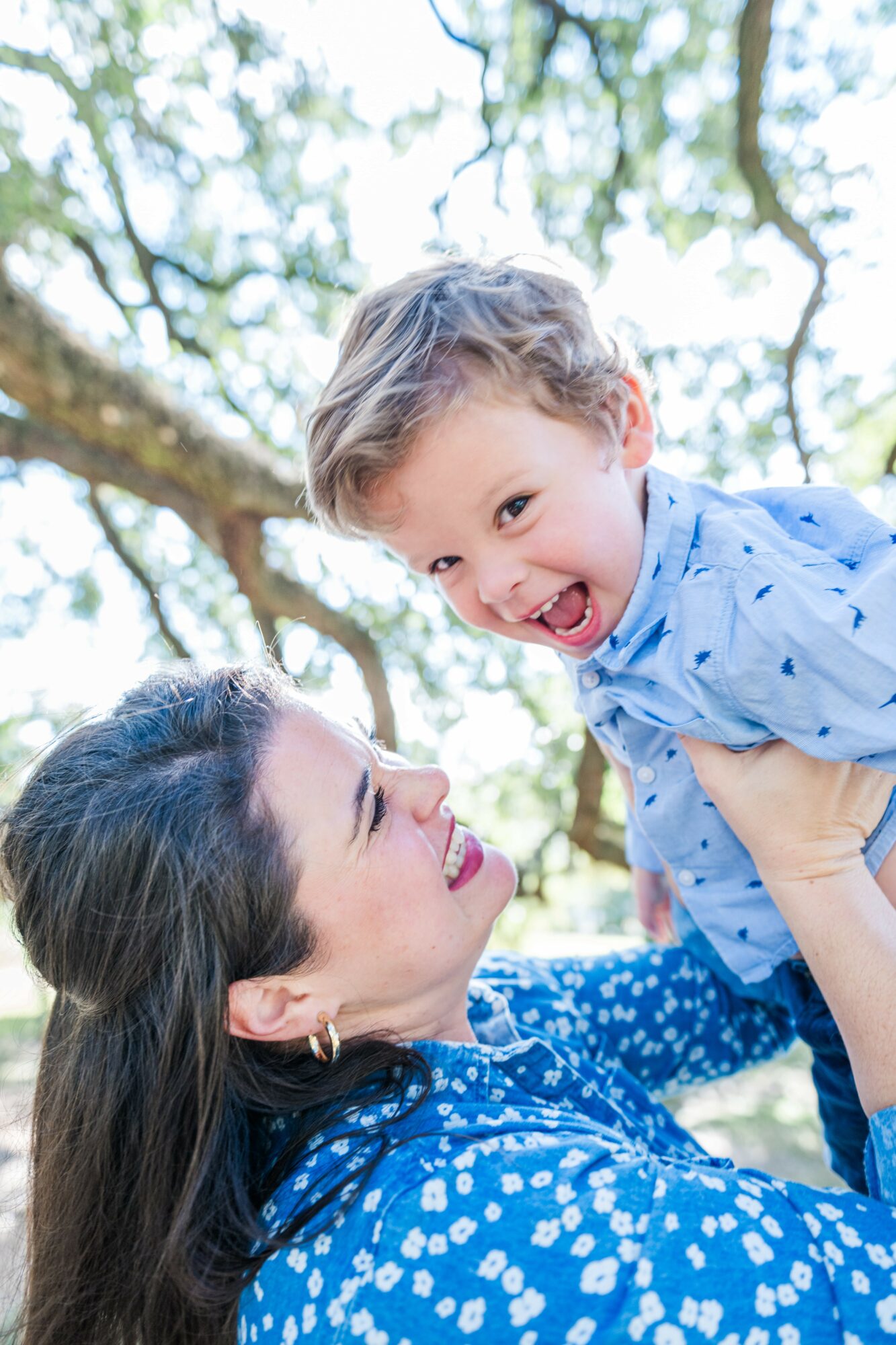 Woman lifting a smiling young boy outdoors with trees in the background.