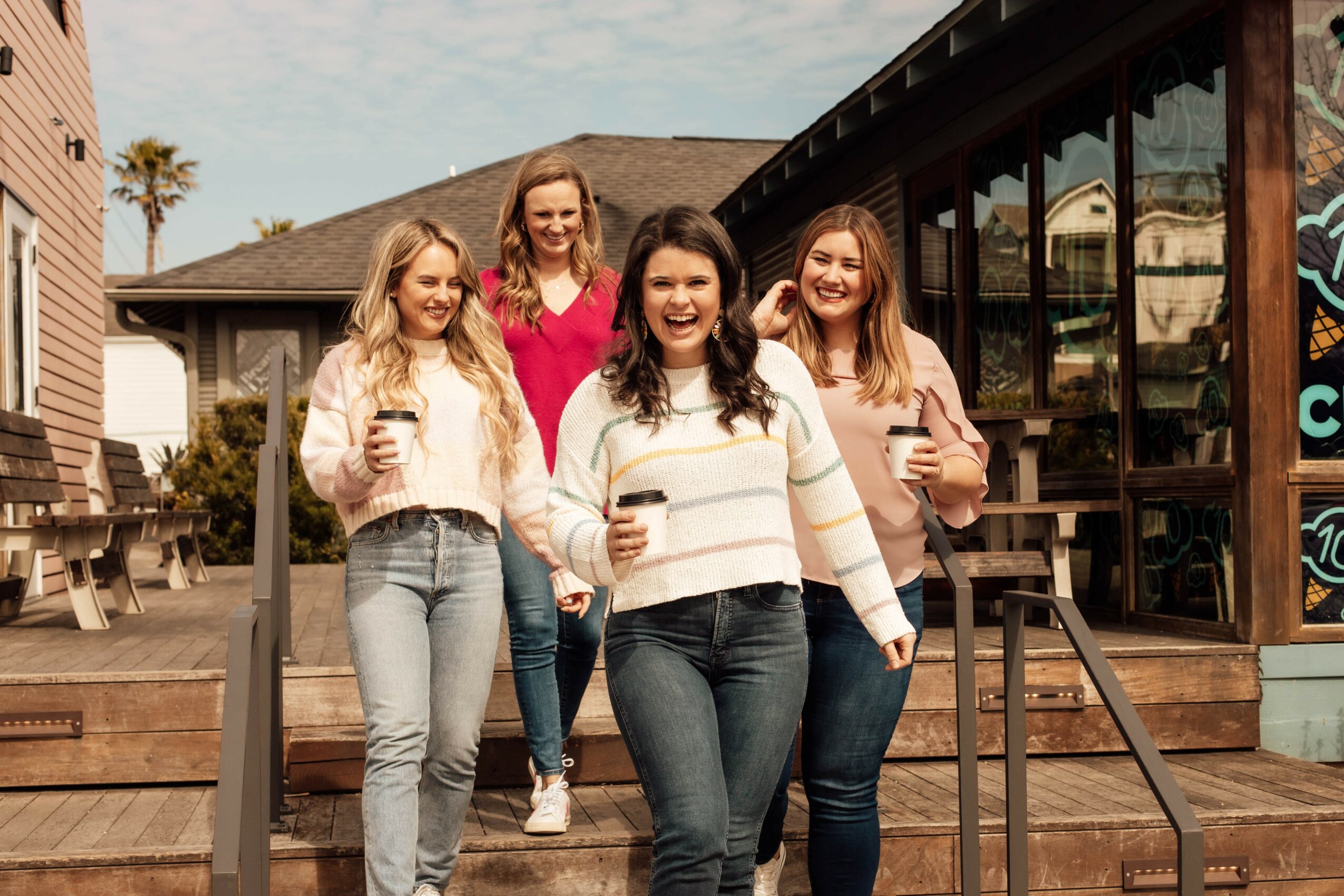 Four women standing on outdoor steps, smiling, holding drinks, in casual clothing, with a building and trees in background.