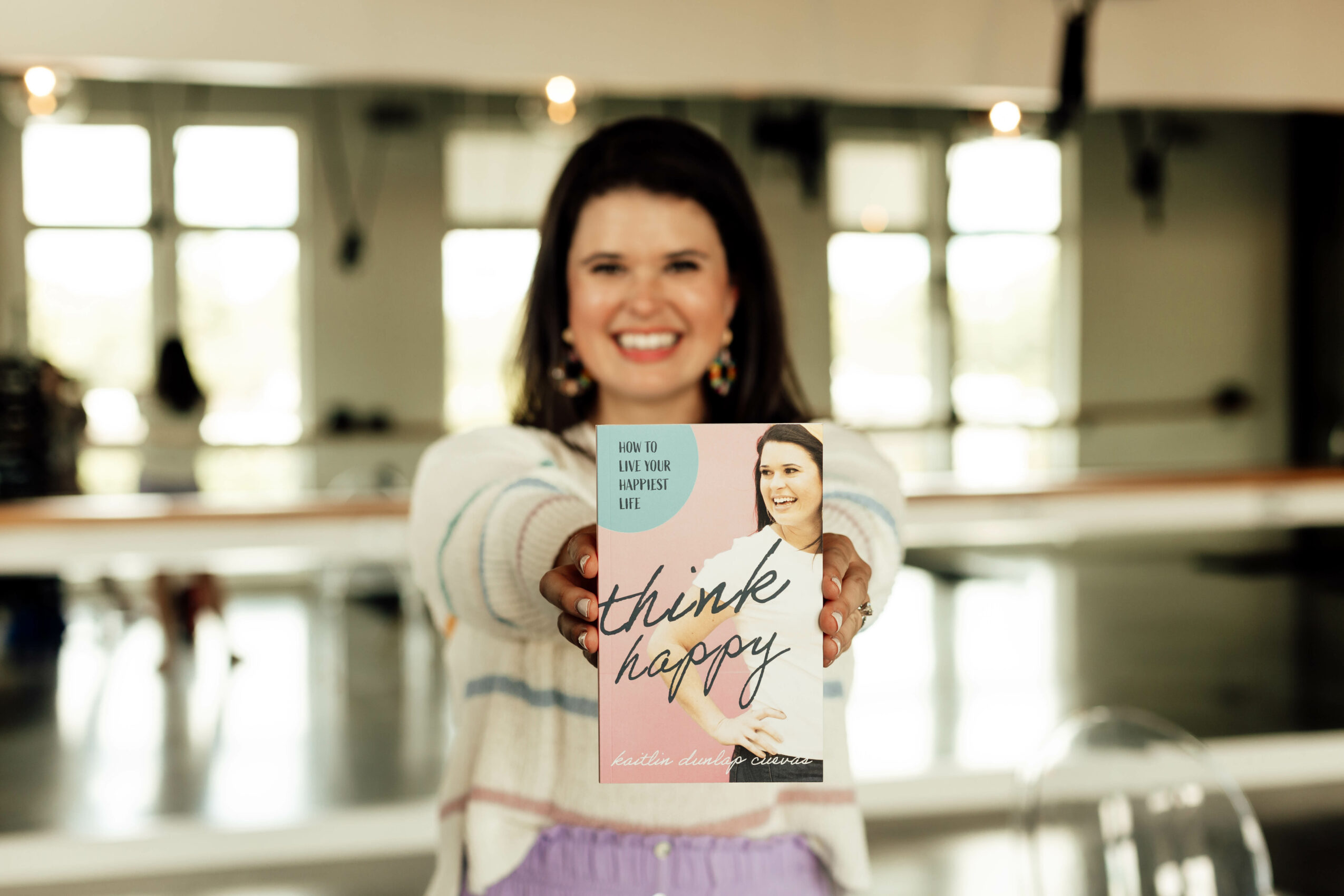Smiling woman holding a book in a bright indoor space with large windows behind her.