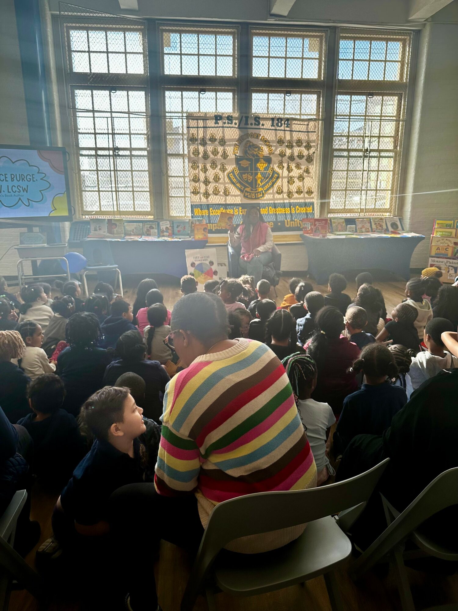 Crowd of children and adults watching a stage with a large banner, inside a spacious room with large windows.