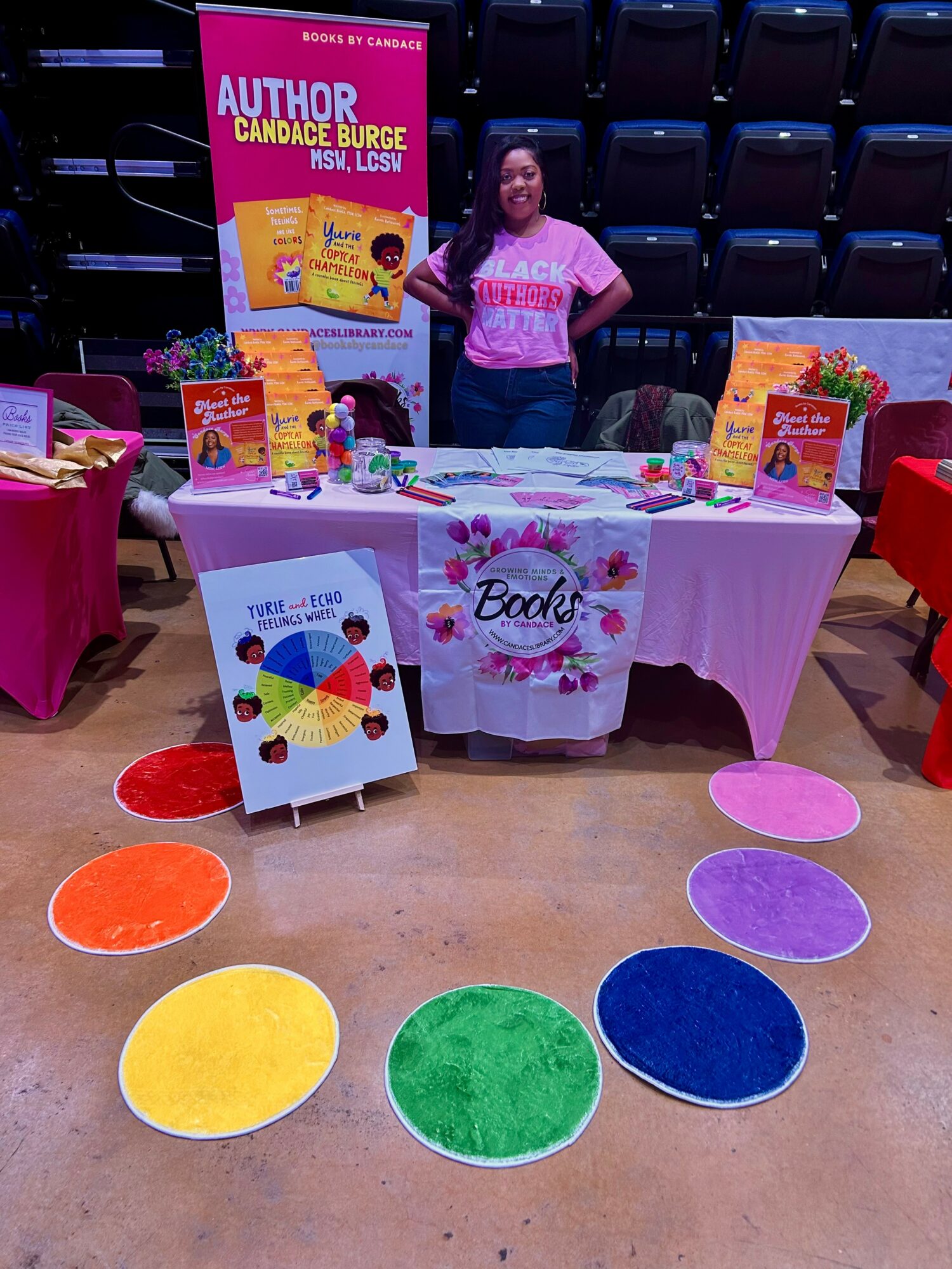 Book signing table with colorful decorations and a person standing behind, in an indoor venue with chairs. Word 'Books' on tablecloth.