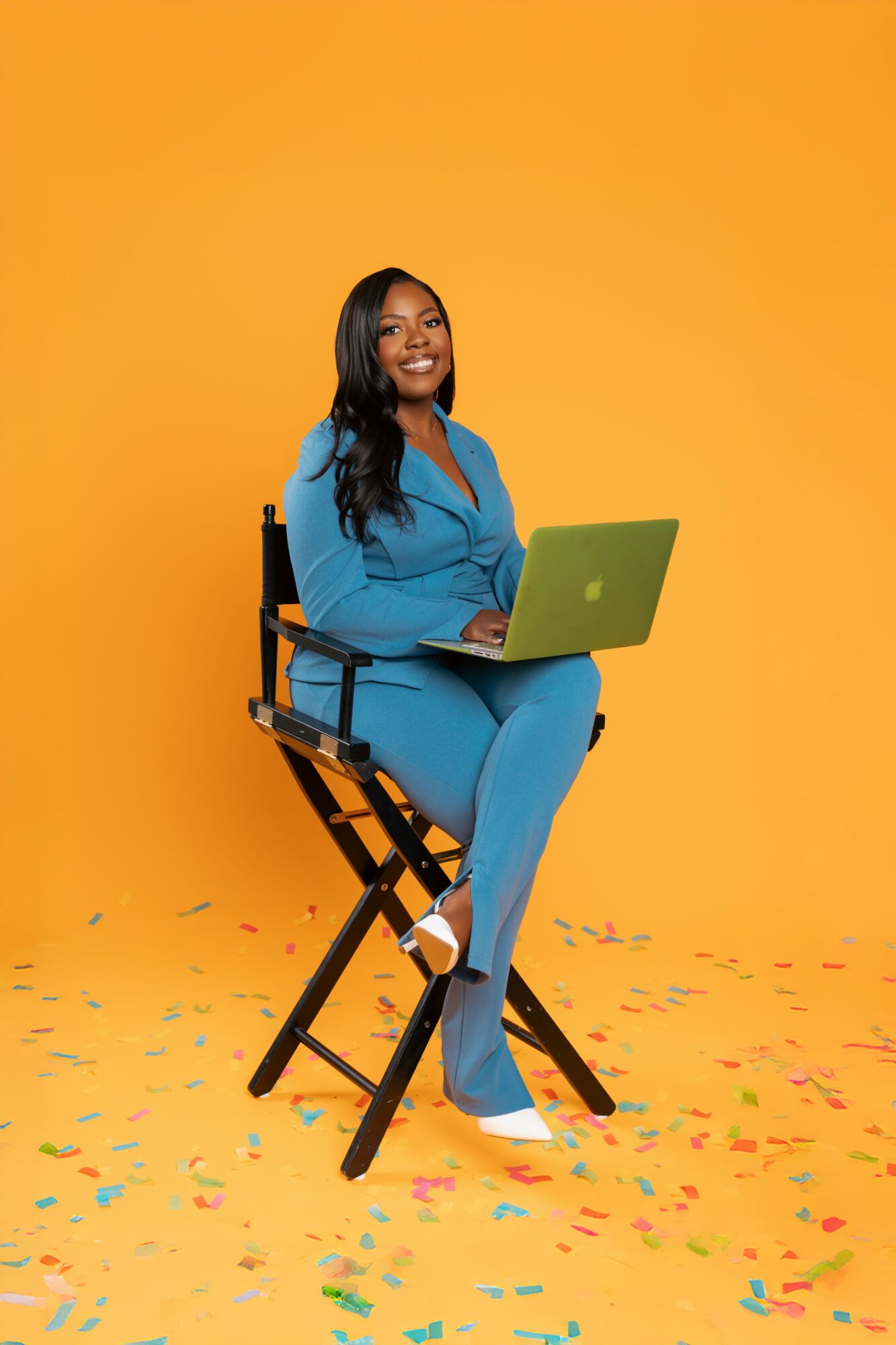 Woman in blue suit sitting on a chair with a laptop, smiling, against a yellow background with confetti on the floor.