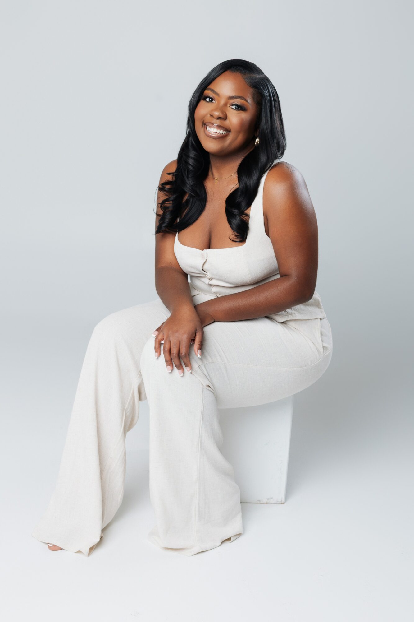 Smiling woman with long dark hair in a white sleeveless outfit, sitting on a white stool against a light background.