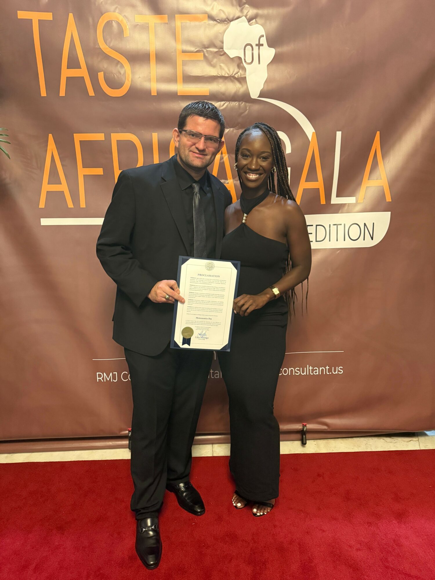 Two people standing in front of a backdrop, one holding a certificate, both smiling, on a red carpet.