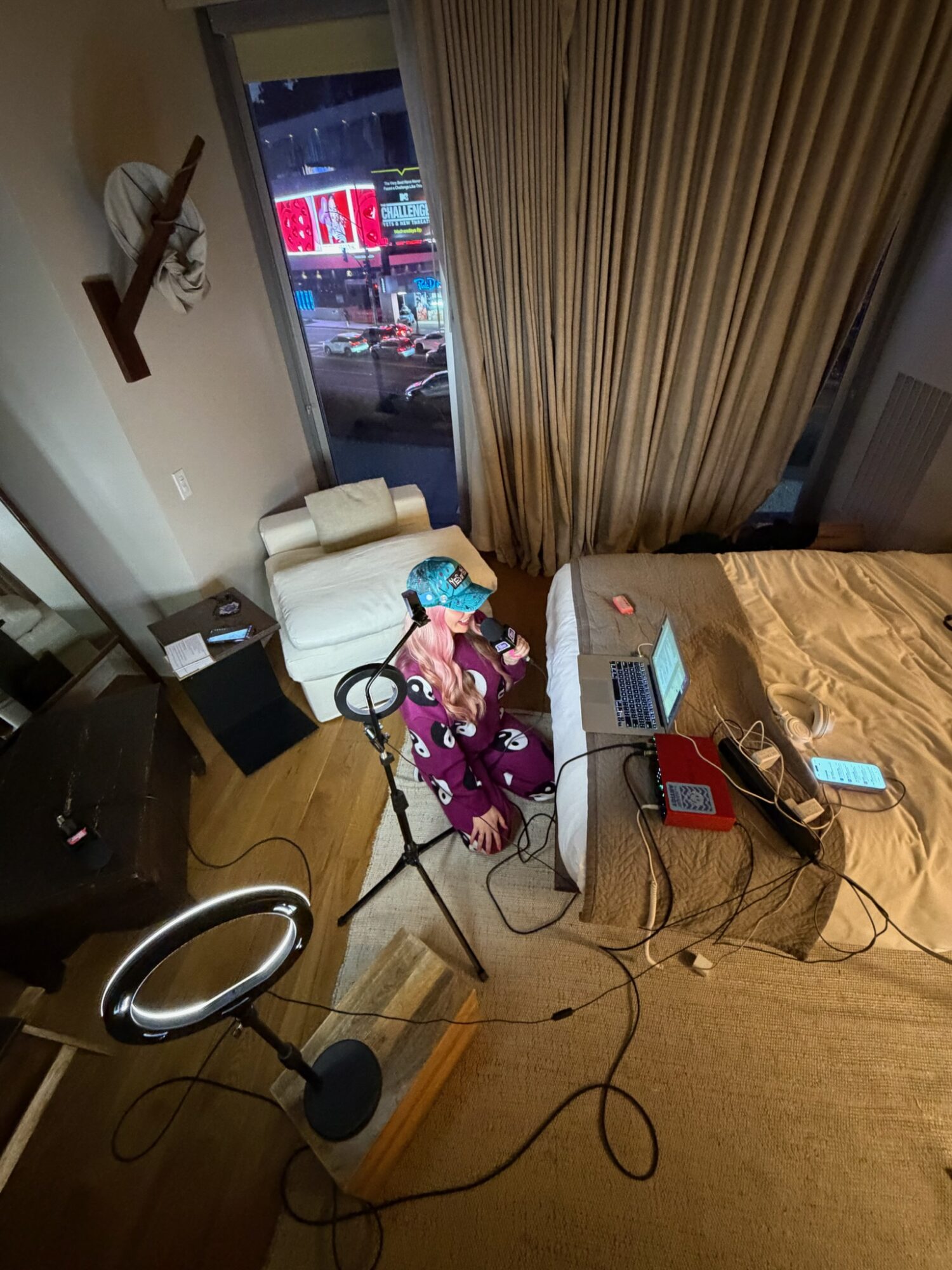 Person sitting on bed with laptop and phone, room with curtains, chair, and window showing city street.