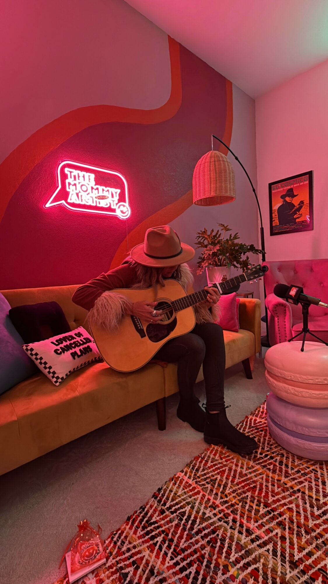 Person wearing a hat playing guitar on a sofa in a colorful room with neon sign and artwork.