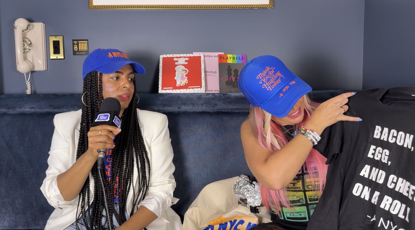 Two women sitting on a couch, one holding a microphone, the other leaning on a bag, with books and a wall phone behind them.